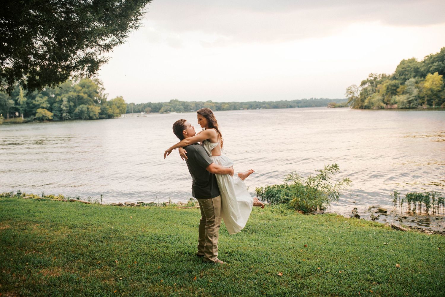 Groom lifting bride by a serene riverside on lush green grass during a romantic outdoor engagement photo session.