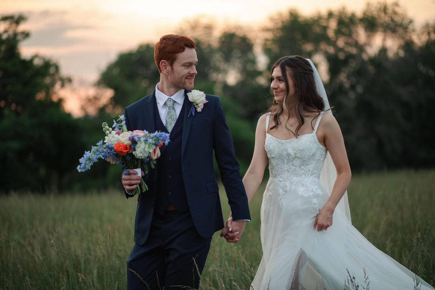 A bride and groom walk hand in hand through a green meadow at sunset, smiling at each other in wedding attire.