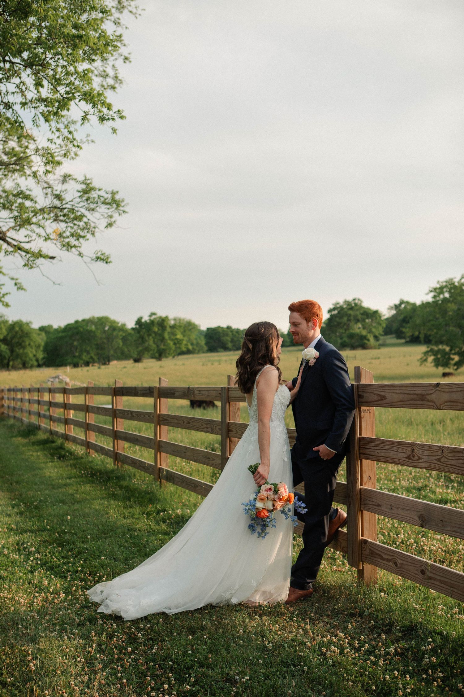 Bride and groom share a romantic moment by a rustic wooden fence in a lush green countryside setting.