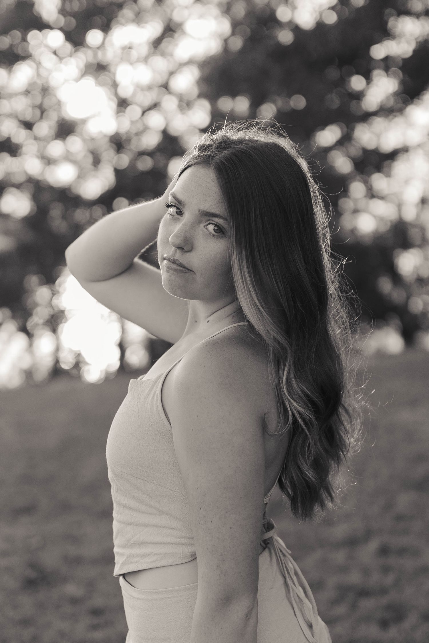 Black and white portrait of a young woman with long wavy hair posing outdoors with bokeh tree background.