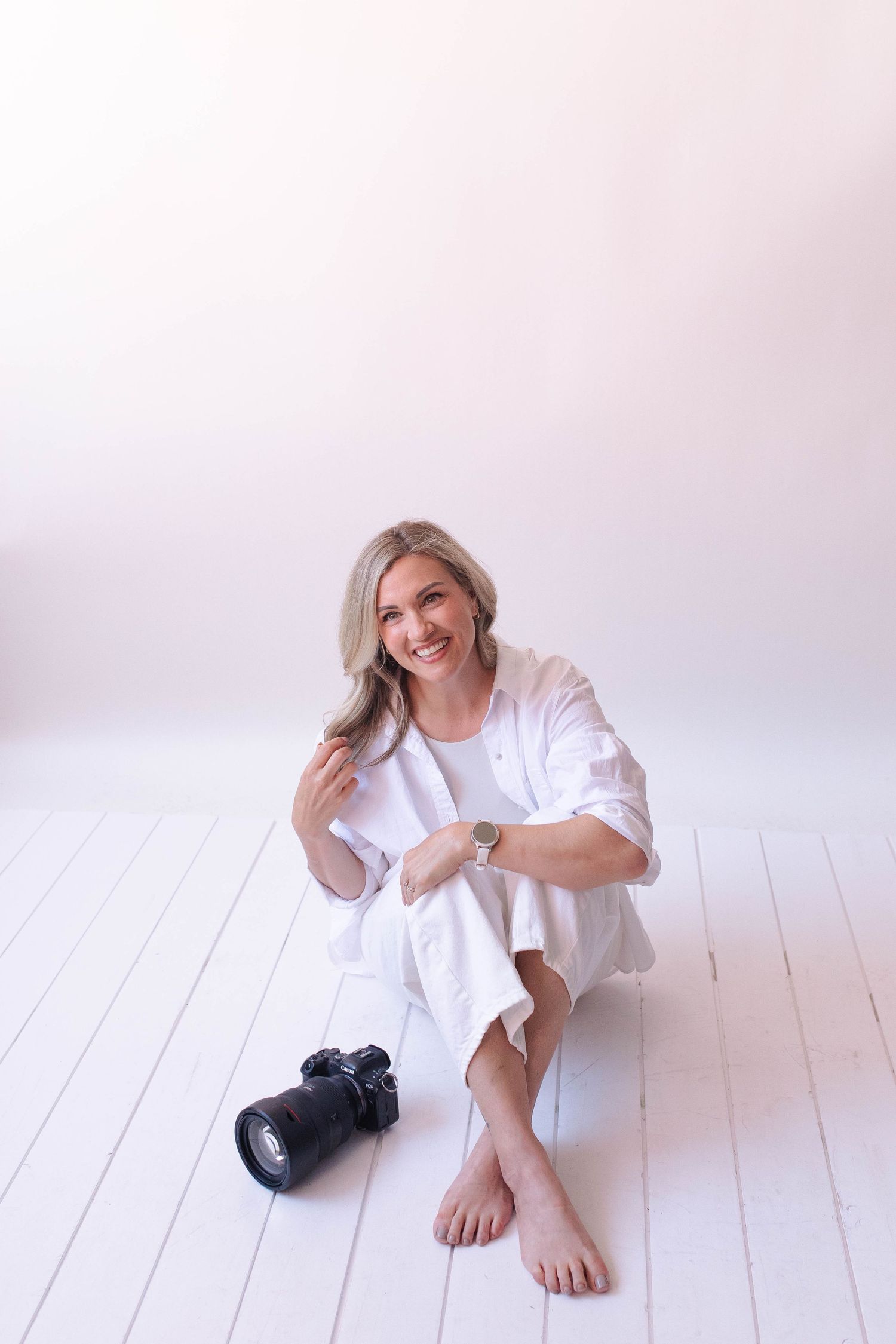 Smiling blonde woman in white outfit sitting barefoot on white floor with DSLR camera beside her.