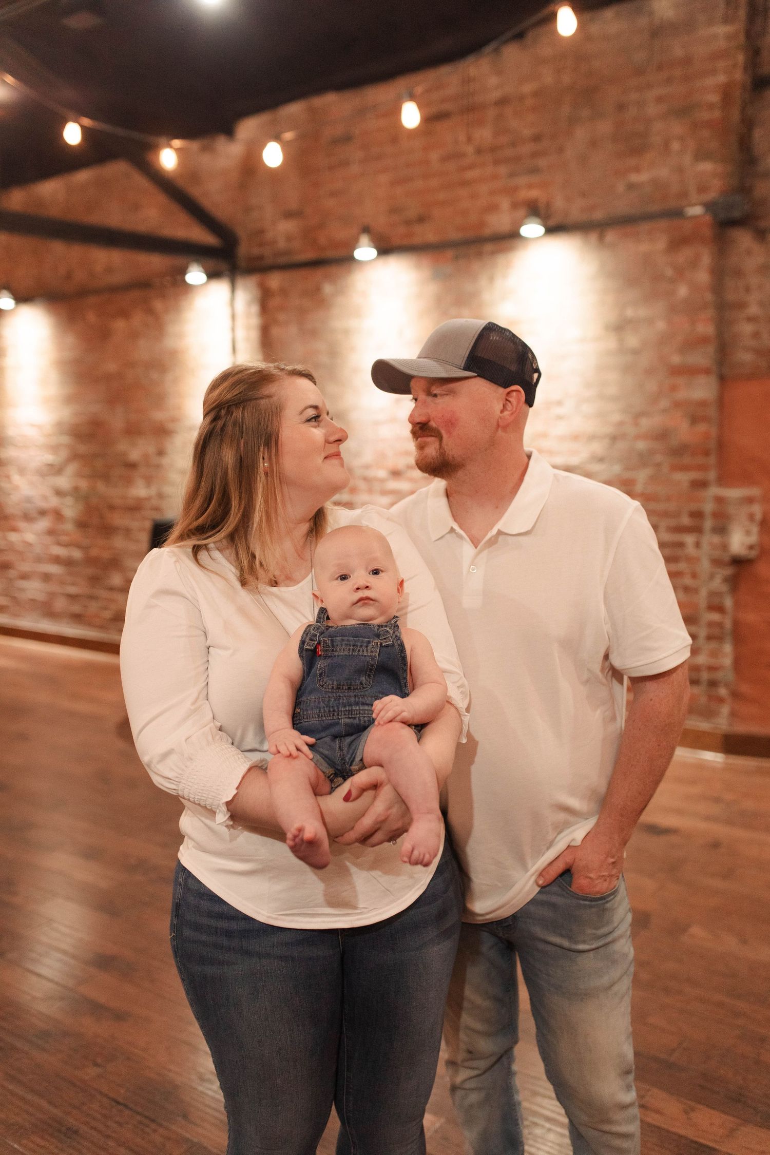 Young couple in white tops holding baby in denim overalls, posing in rustic brick venue with string lights.
