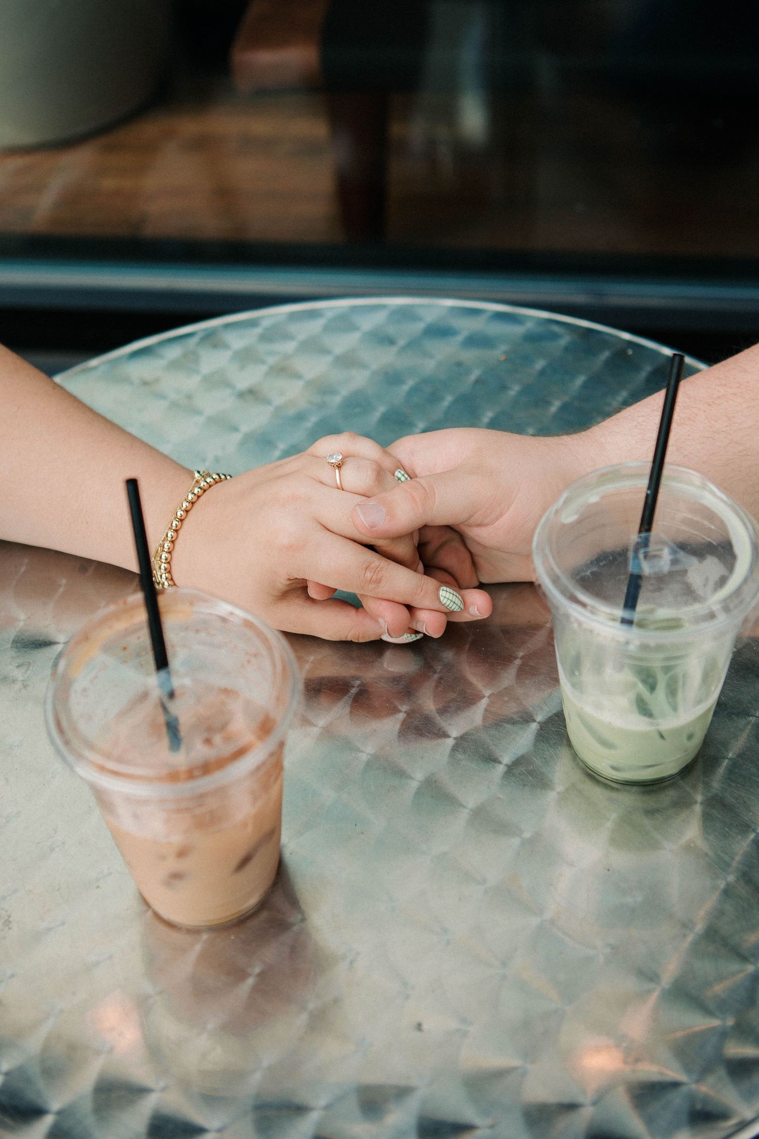 Two people holding hands on a glass table with iced coffee and matcha latte drinks with black straws.