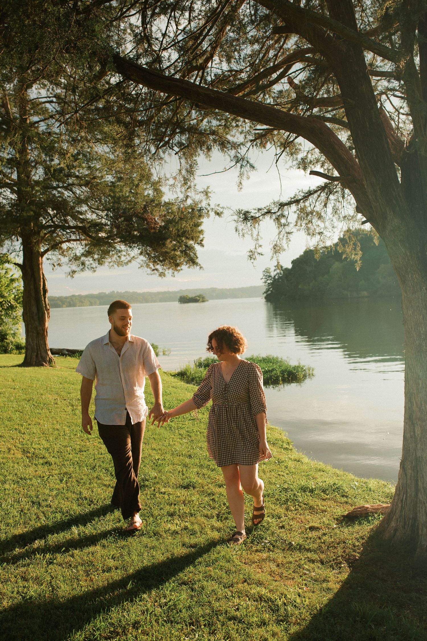 Couple holding hands walking by a serene lake at golden hour, surrounded by lush green trees and grass.