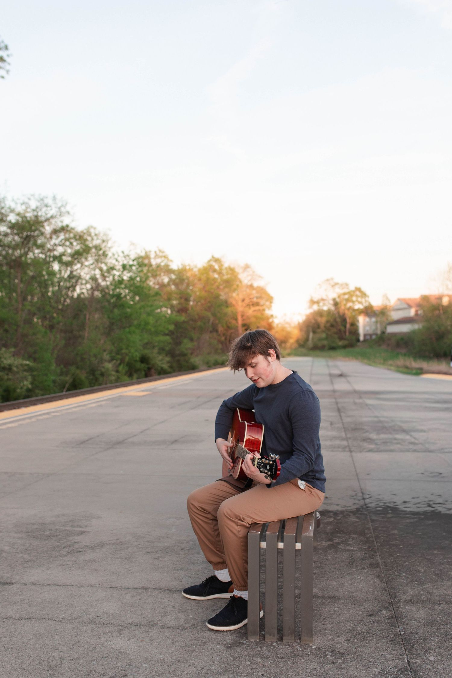 Young man sitting outdoors playing acoustic guitar at sunset on an empty road surrounded by trees.