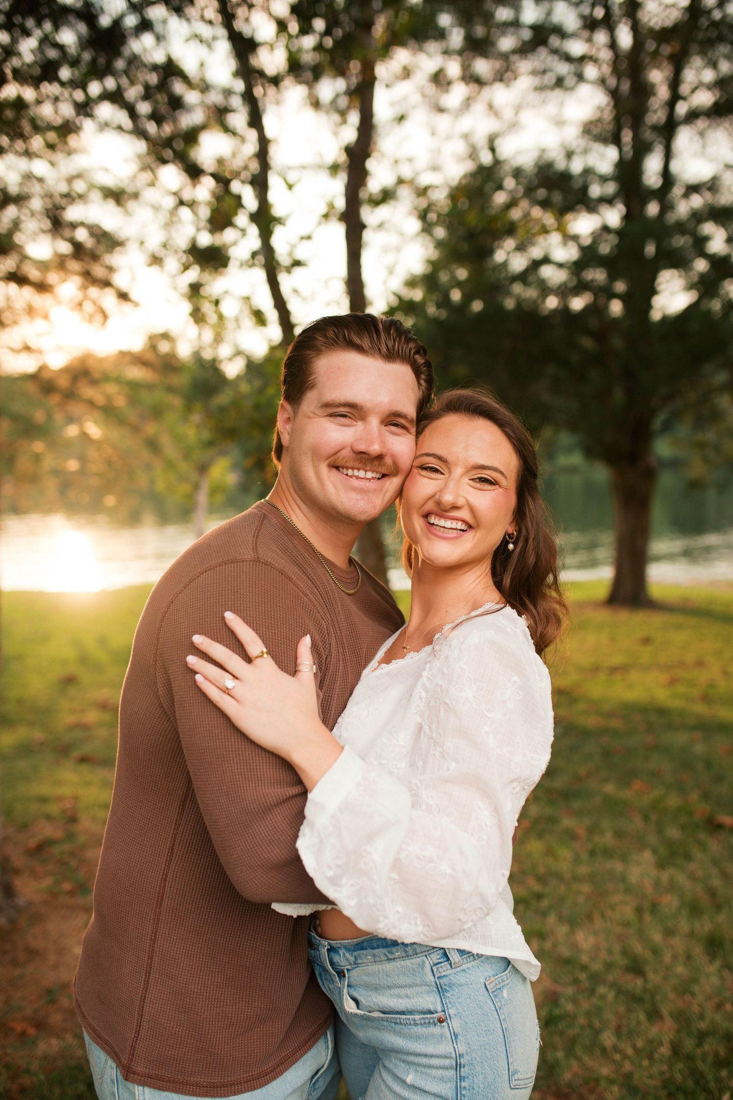 Happy couple embracing at golden hour sunset in a park, woman wearing white lace top and jeans, man in brown shirt.