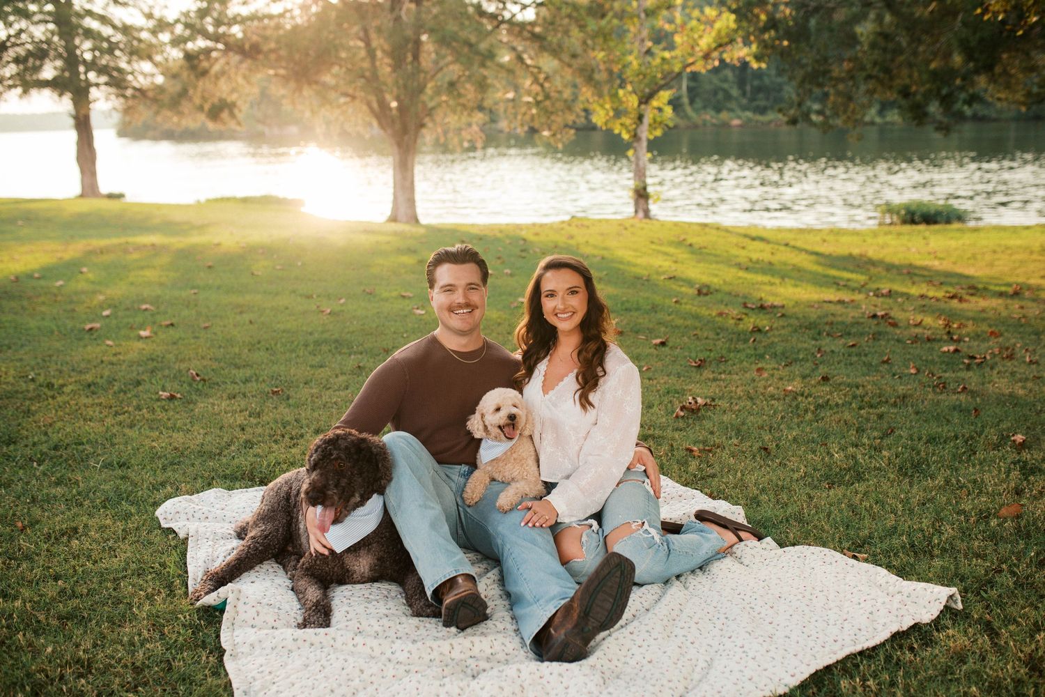 Couple sitting on white blanket in sunny park with two dogs, golden hour light, trees and lake in background.