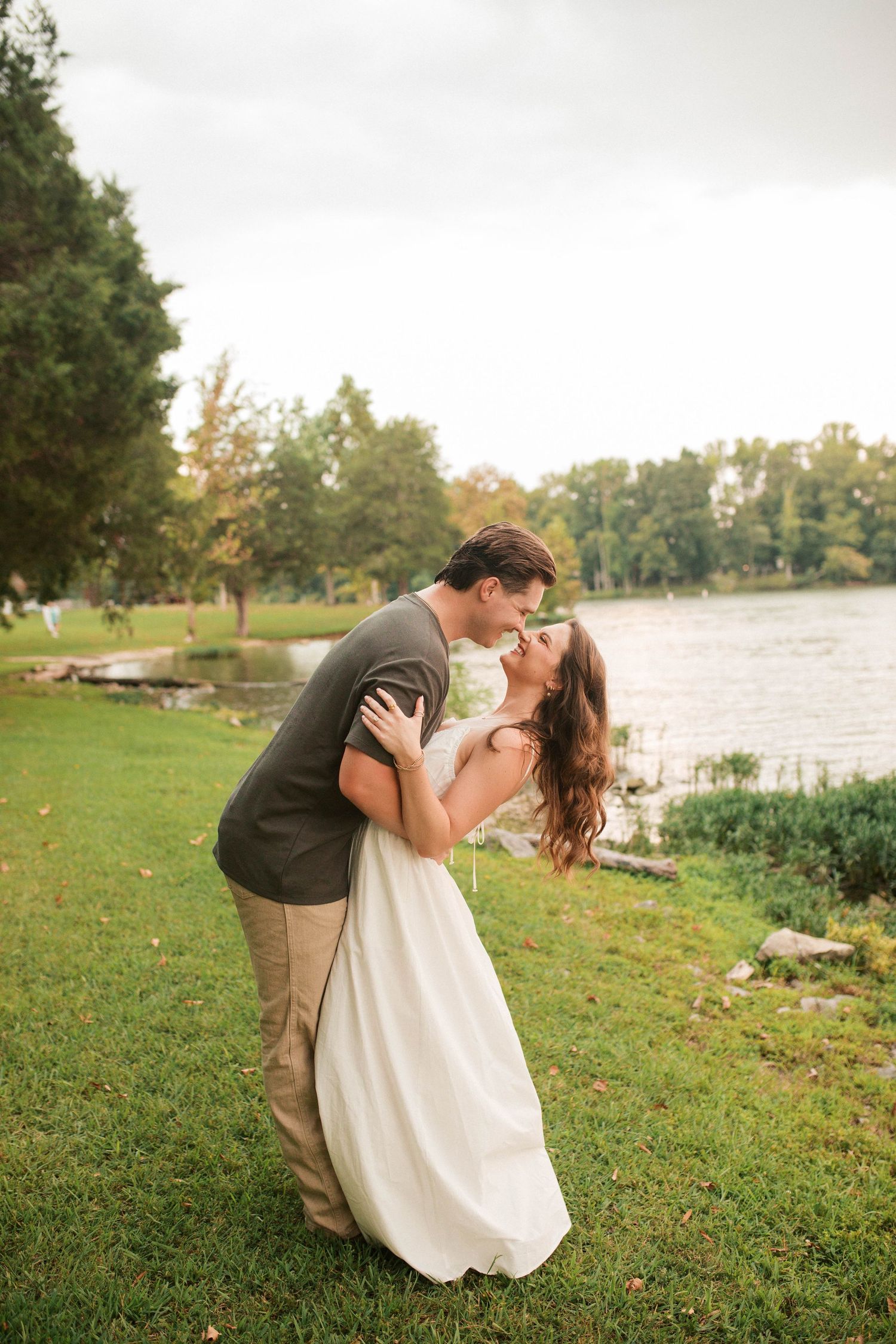 A couple shares a romantic dip kiss near a lakeside park, the bride in a white gown and groom in a gray suit.