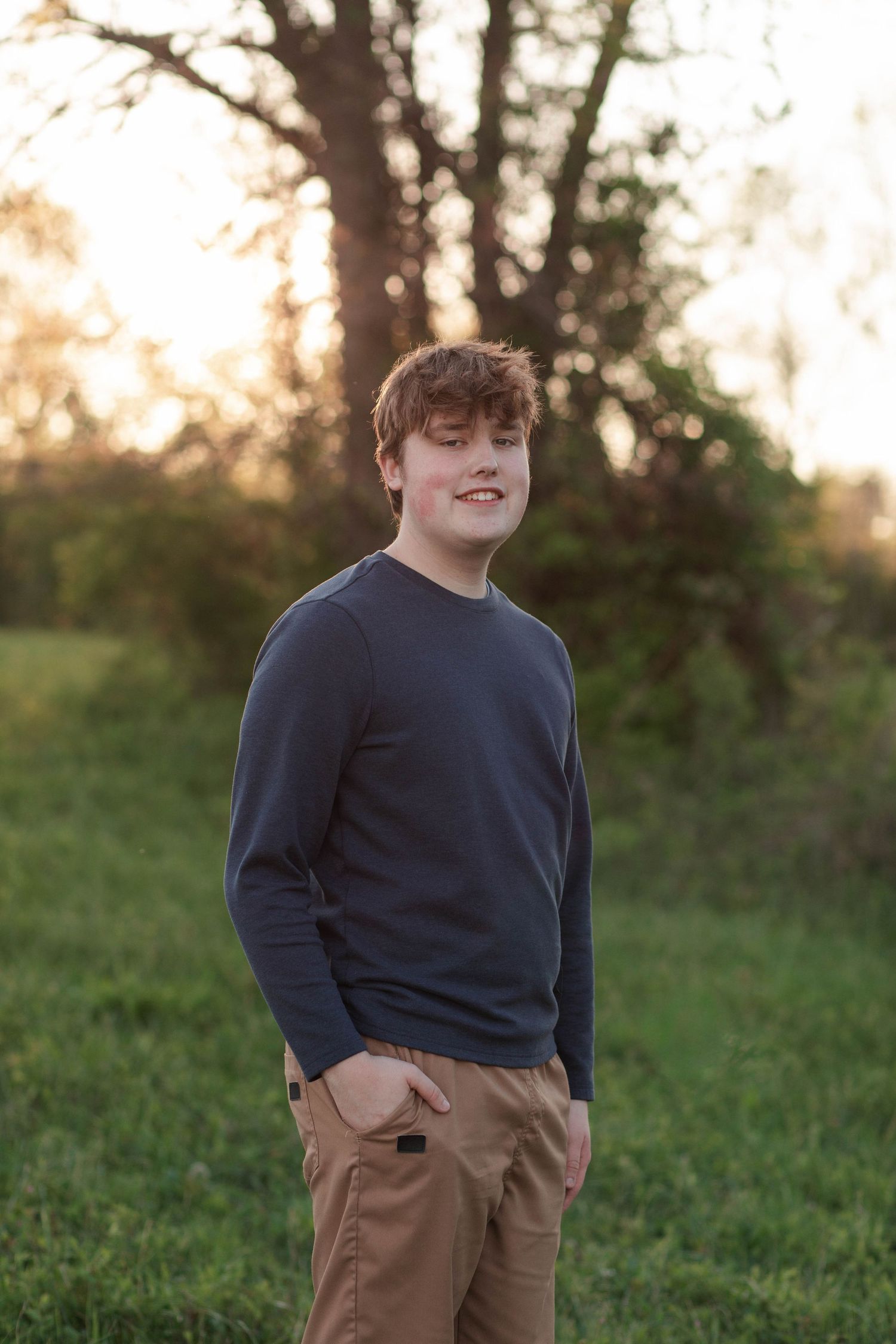 Young man with red hair in navy sweater and khaki pants smiling outdoors in green field at golden hour.