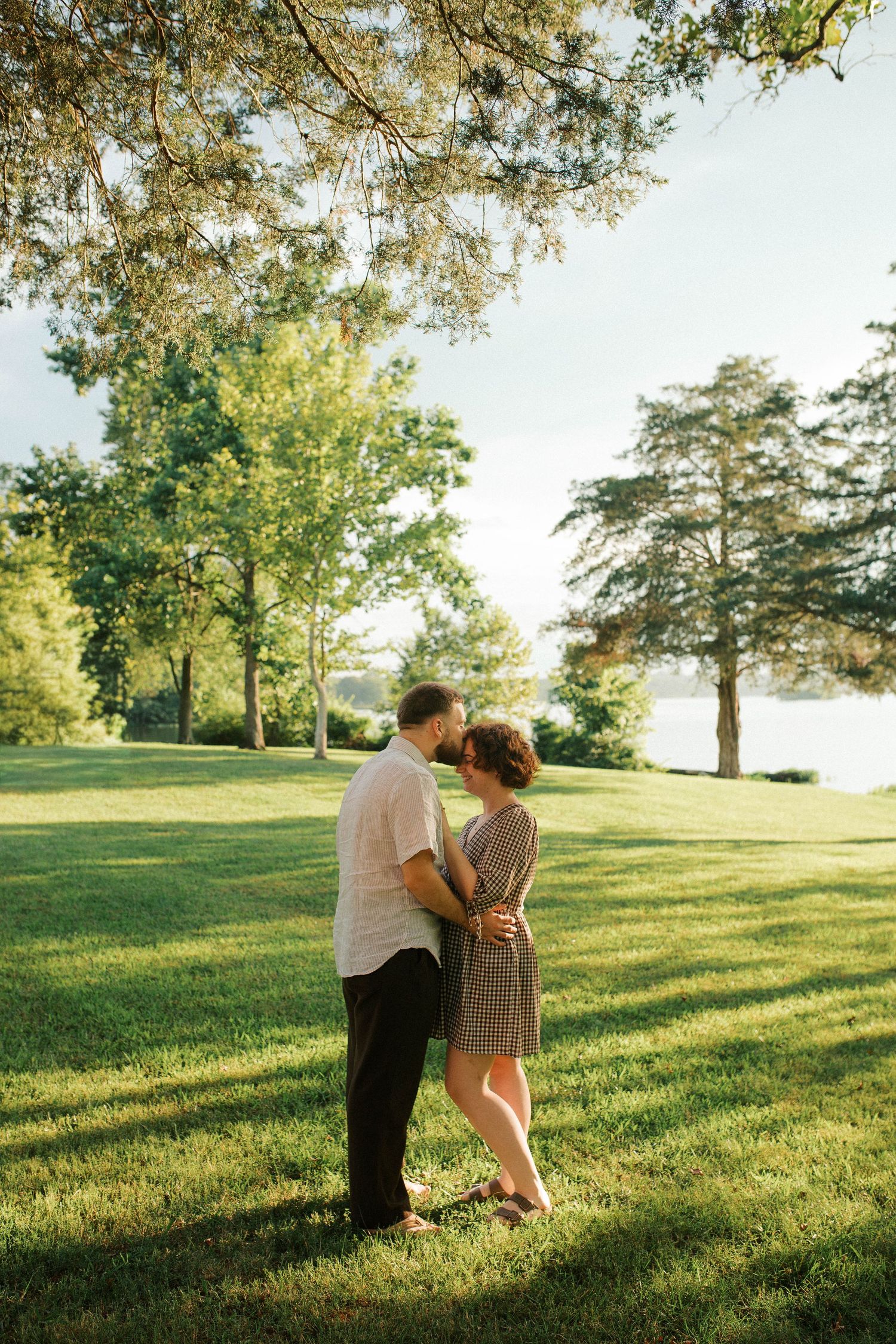 Couple embracing on lush green grass in a sunlit park with tall trees and a lake in the background.