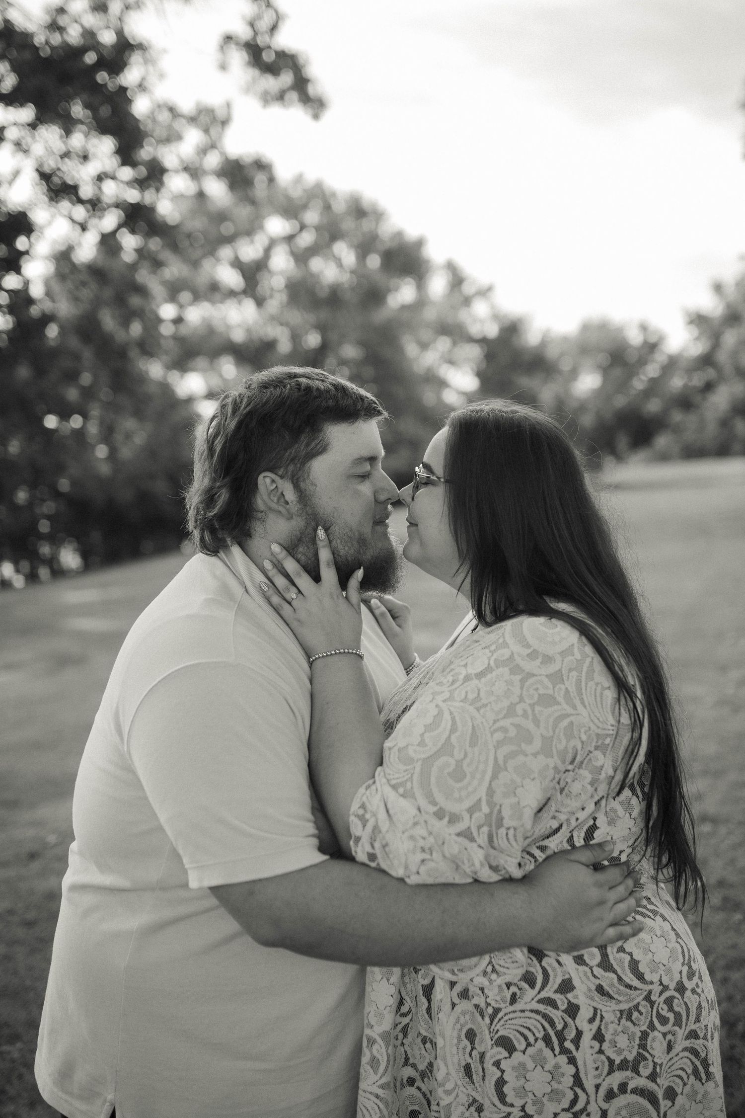 Black and white photo of a couple sharing a romantic kiss outdoors in a park setting.