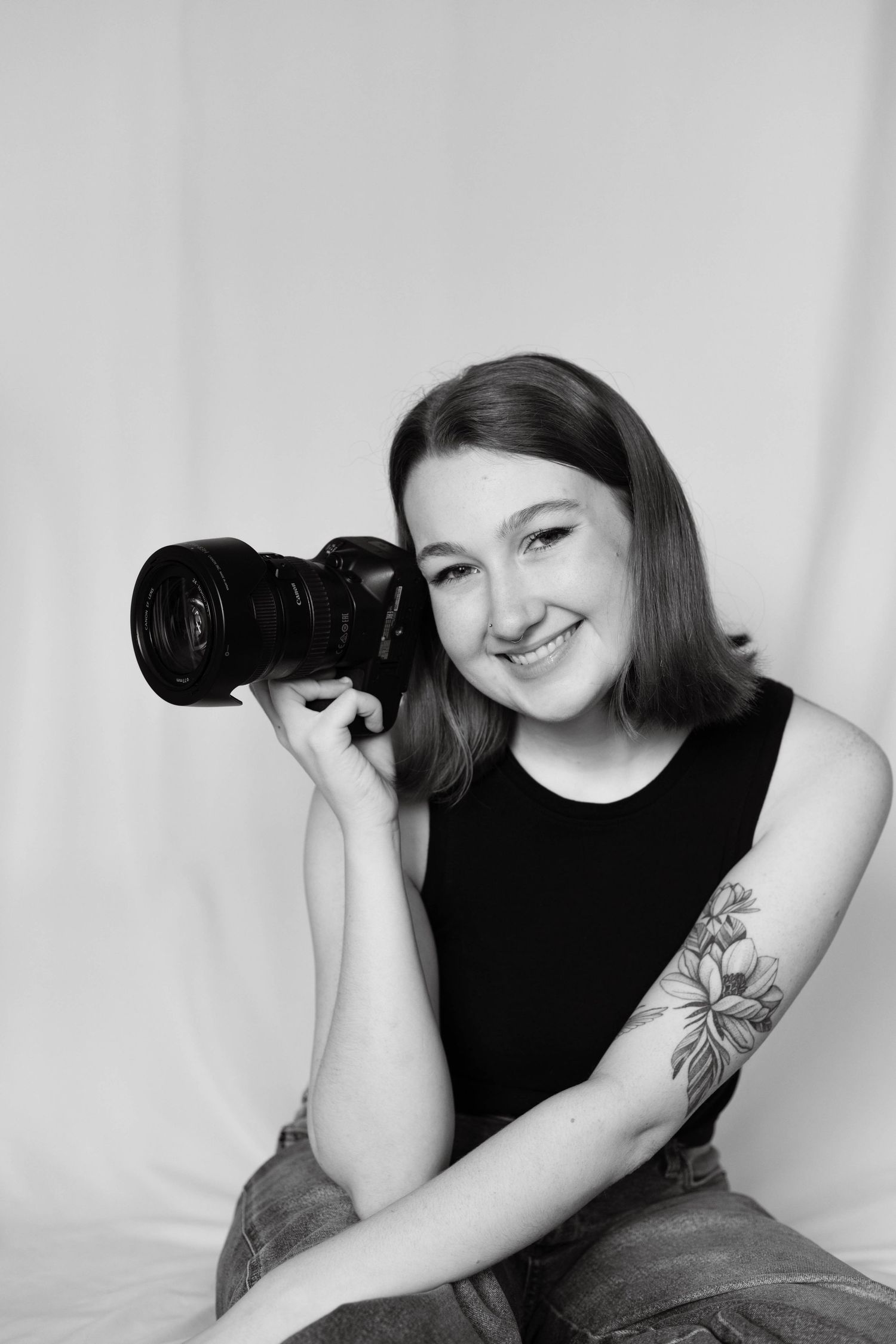 A smiling young woman with a floral arm tattoo holds a DSLR camera in a black and white portrait.