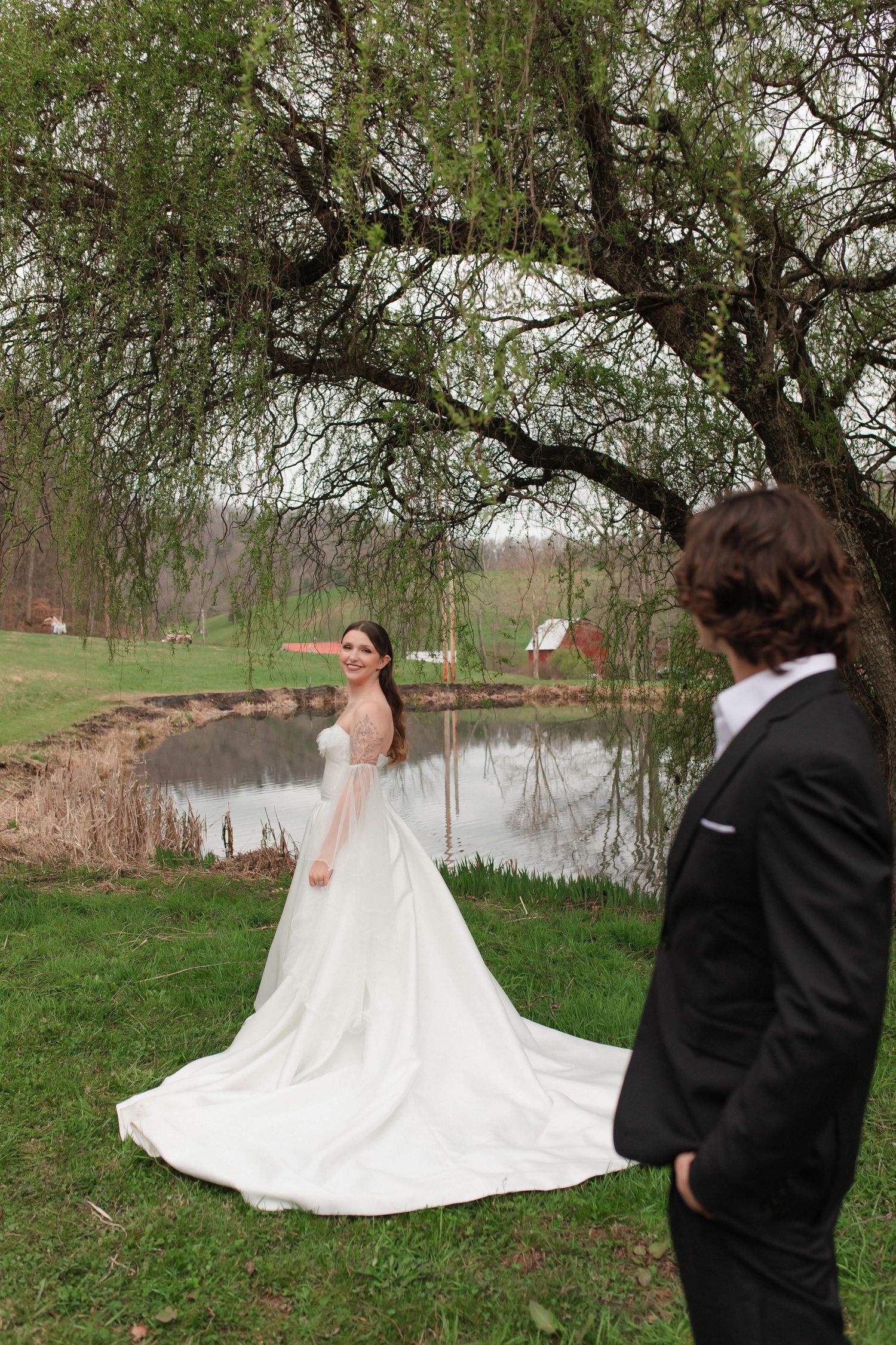 Bride in white gown with long train stands by a pond under a willow tree as groom watches in black suit after eloping in Nashville.