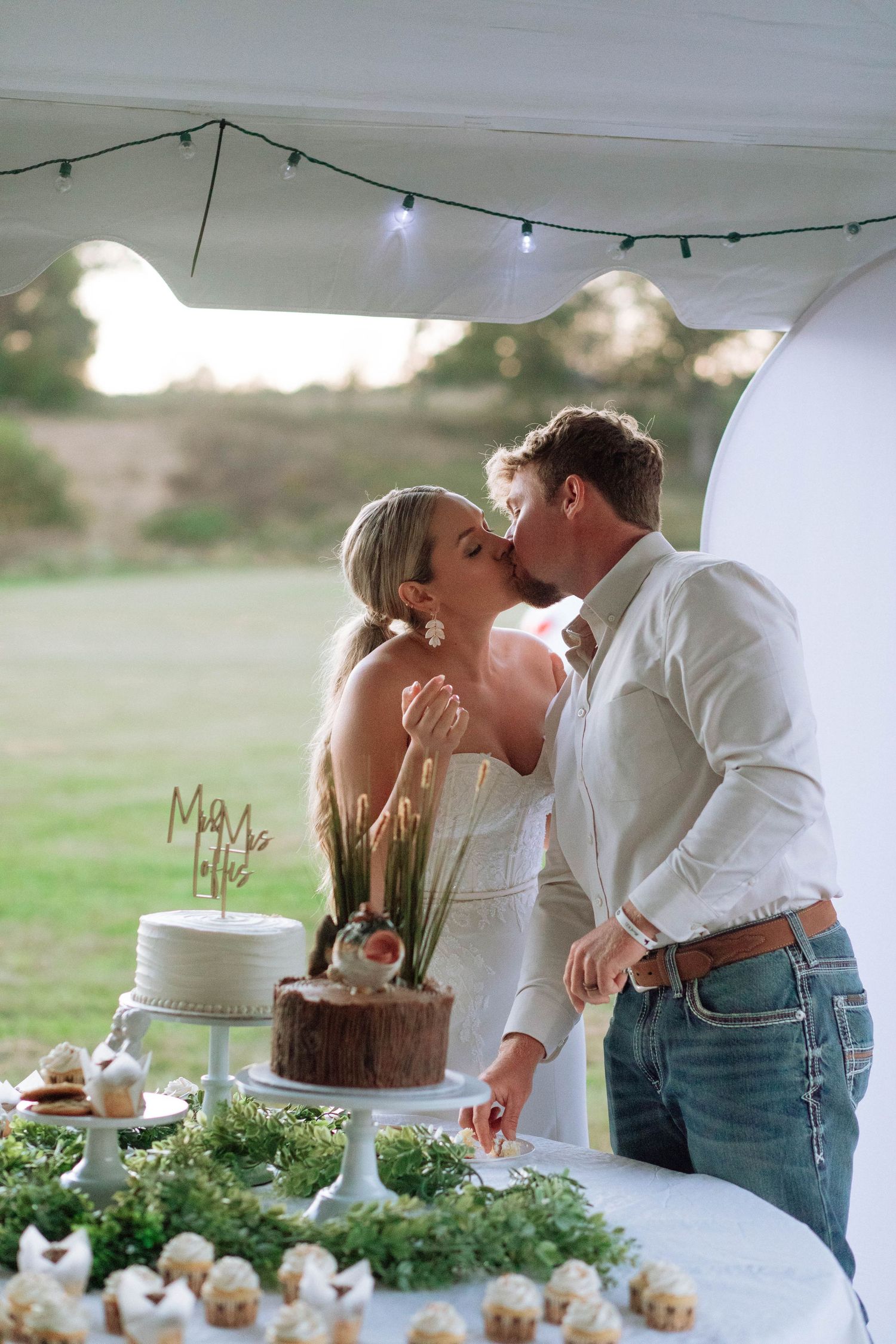 Nashville newlyweds share a kiss while cutting their rustic wedding cake at an outdoor reception at sunset.