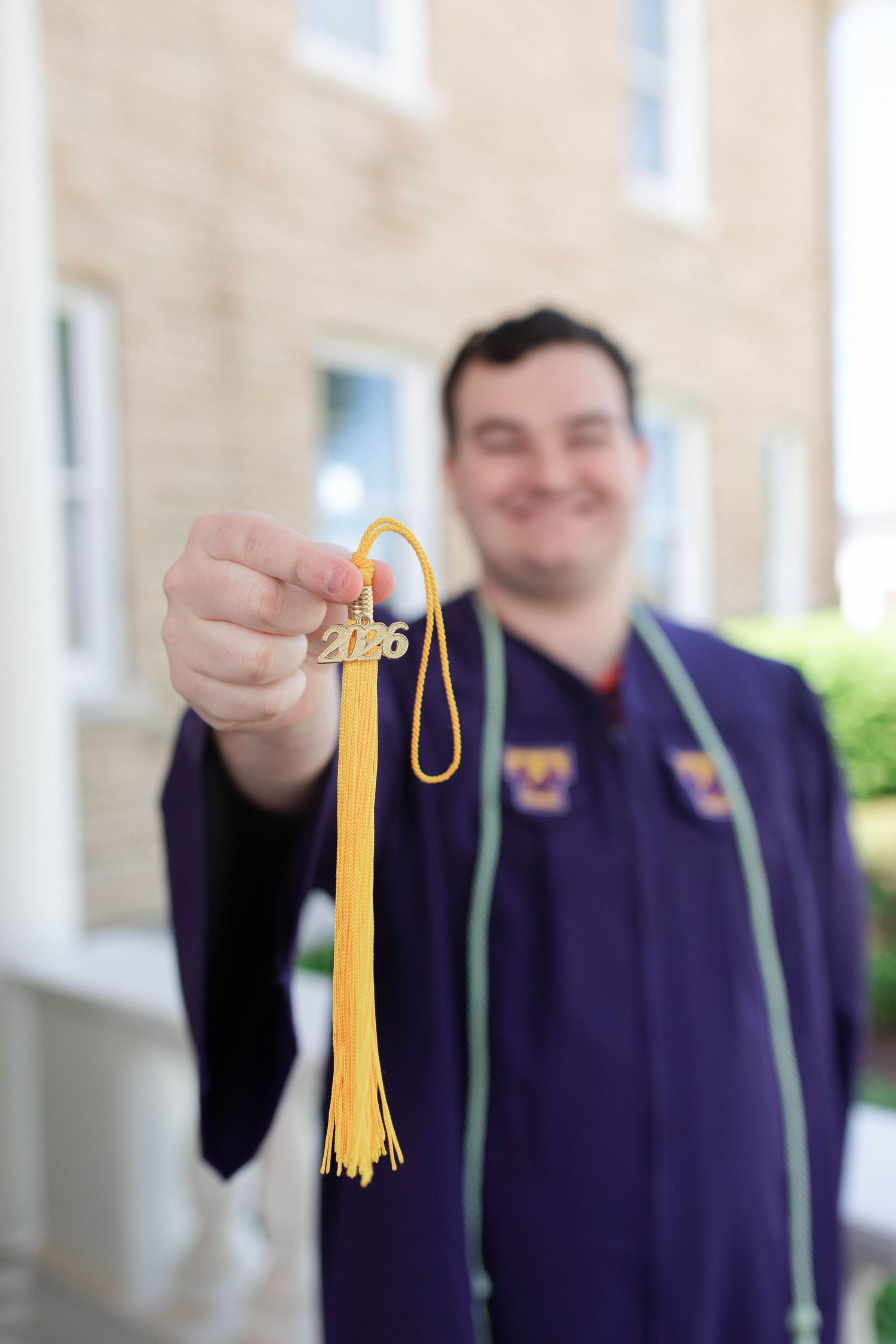 Graduate in purple gown holds up a gold 2026 graduation tassel charm outside a brick building.