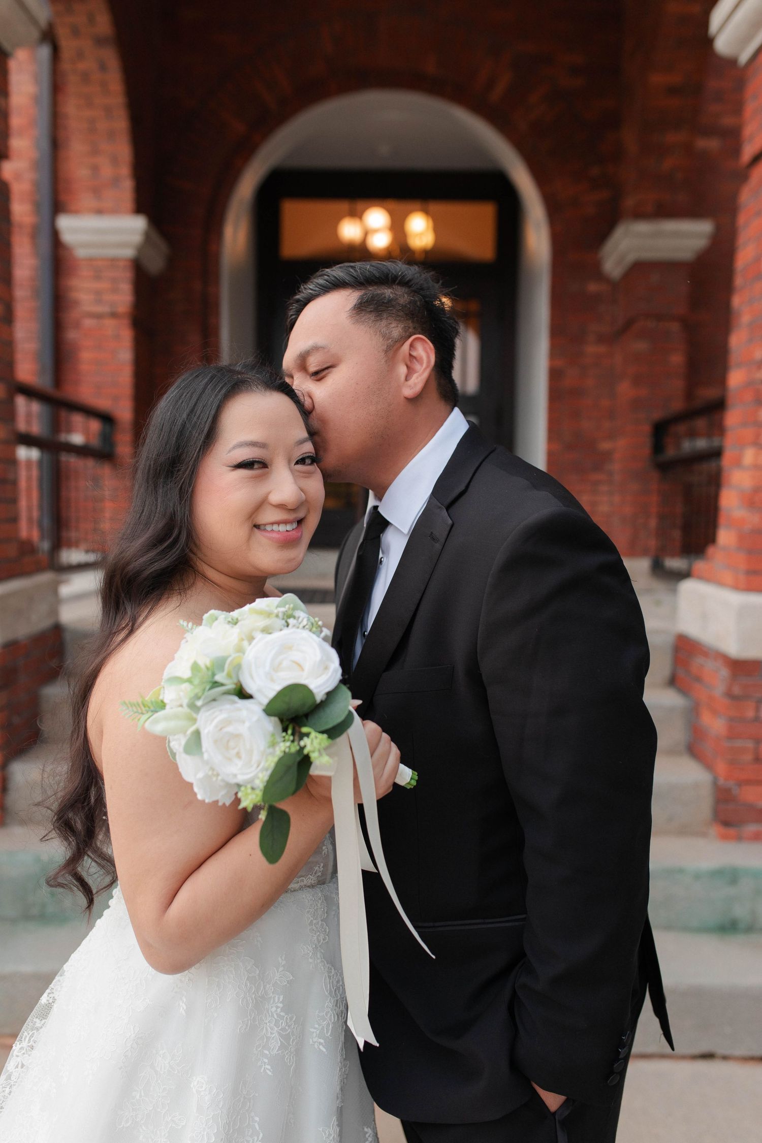 A groom in a black suit kisses his bride on the cheek as she smiles holding white roses outside a brick building.