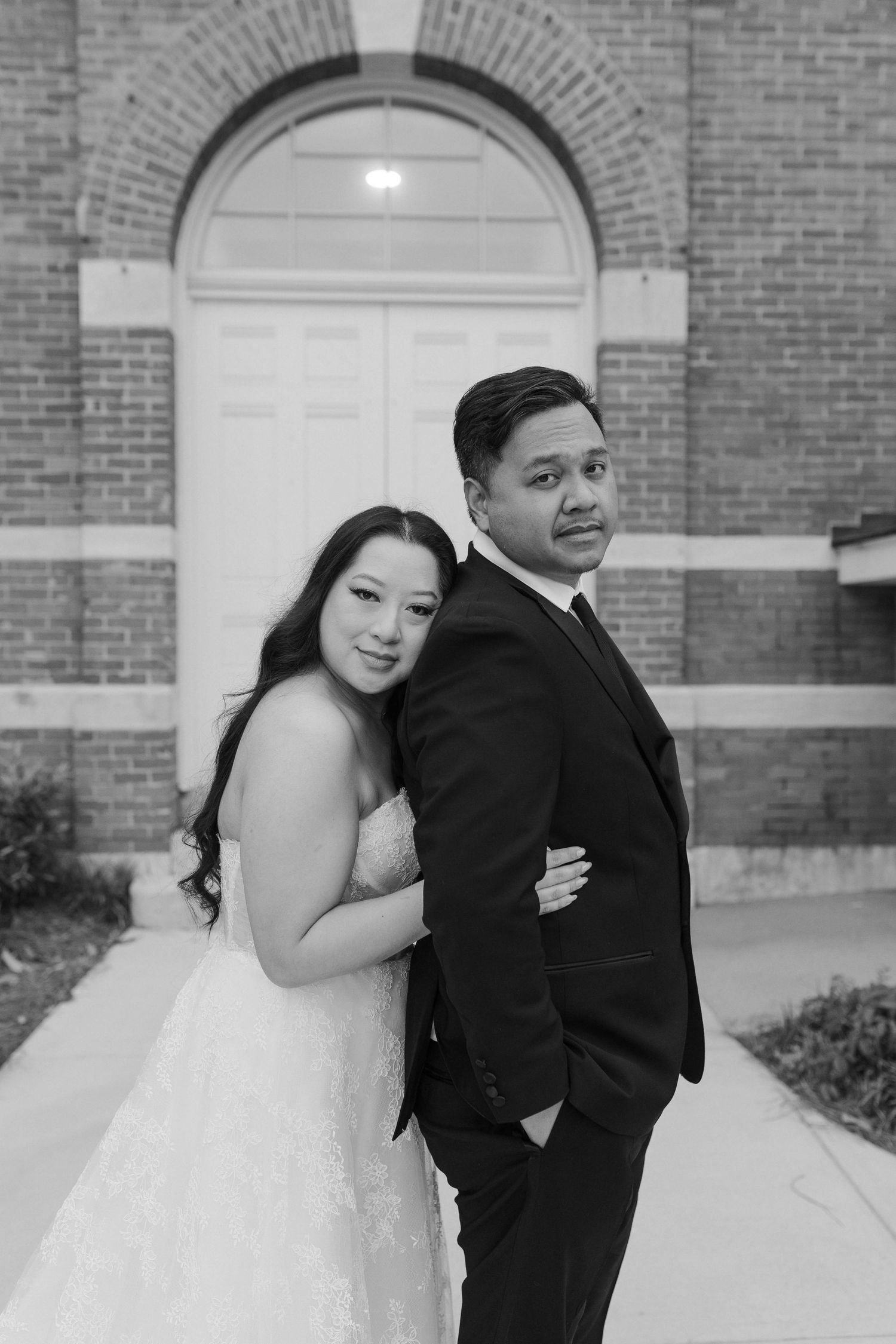 Black and white wedding photo of bride and groom posing back-to-back outside a brick church.