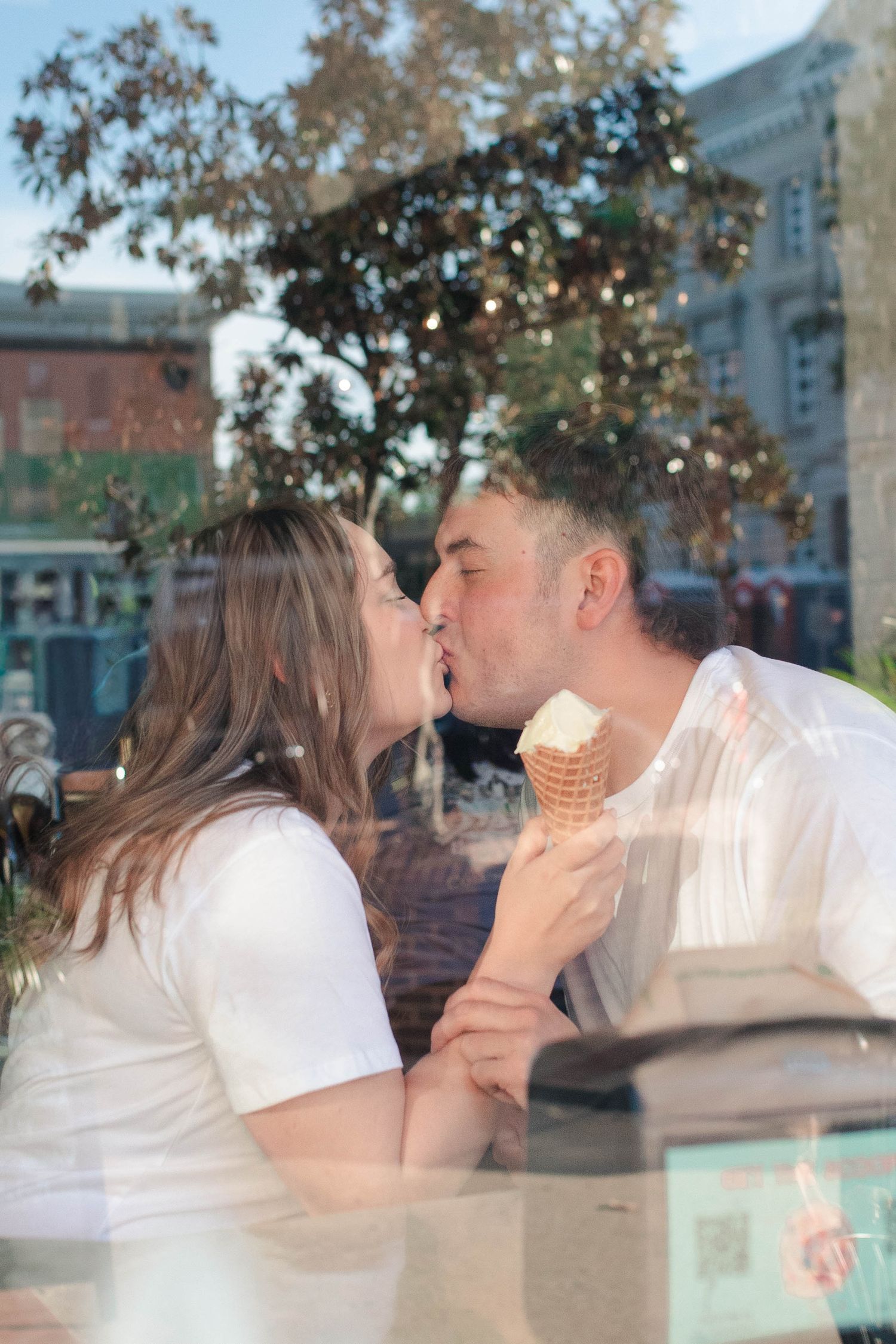 Couple sharing a romantic kiss through a window at an ice cream shop, holding a waffle cone.