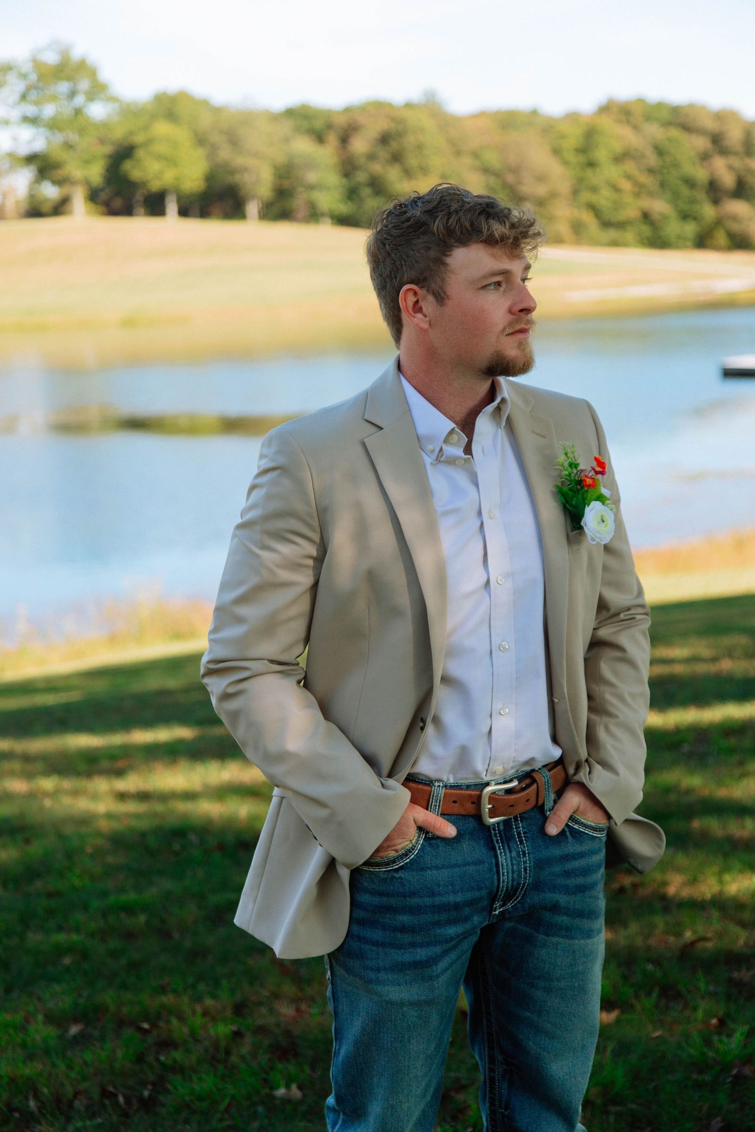 Groom in tan blazer and jeans with floral boutonniere standing near a lake with trees in background before eloping in Nashville.