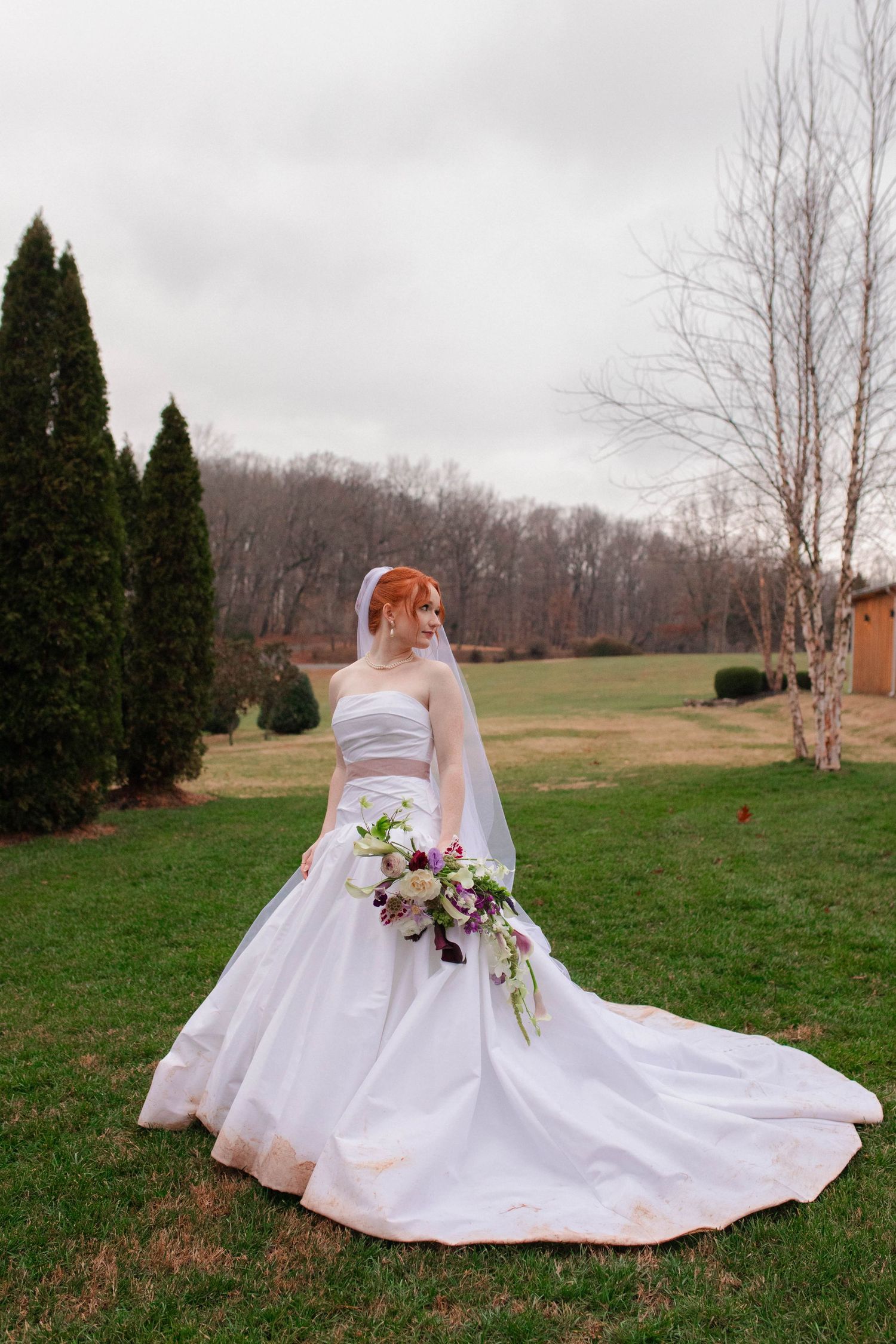 Red-haired bride in strapless white ballgown with veil holds purple and white bouquet on green lawn after micro wedding in Nashville.