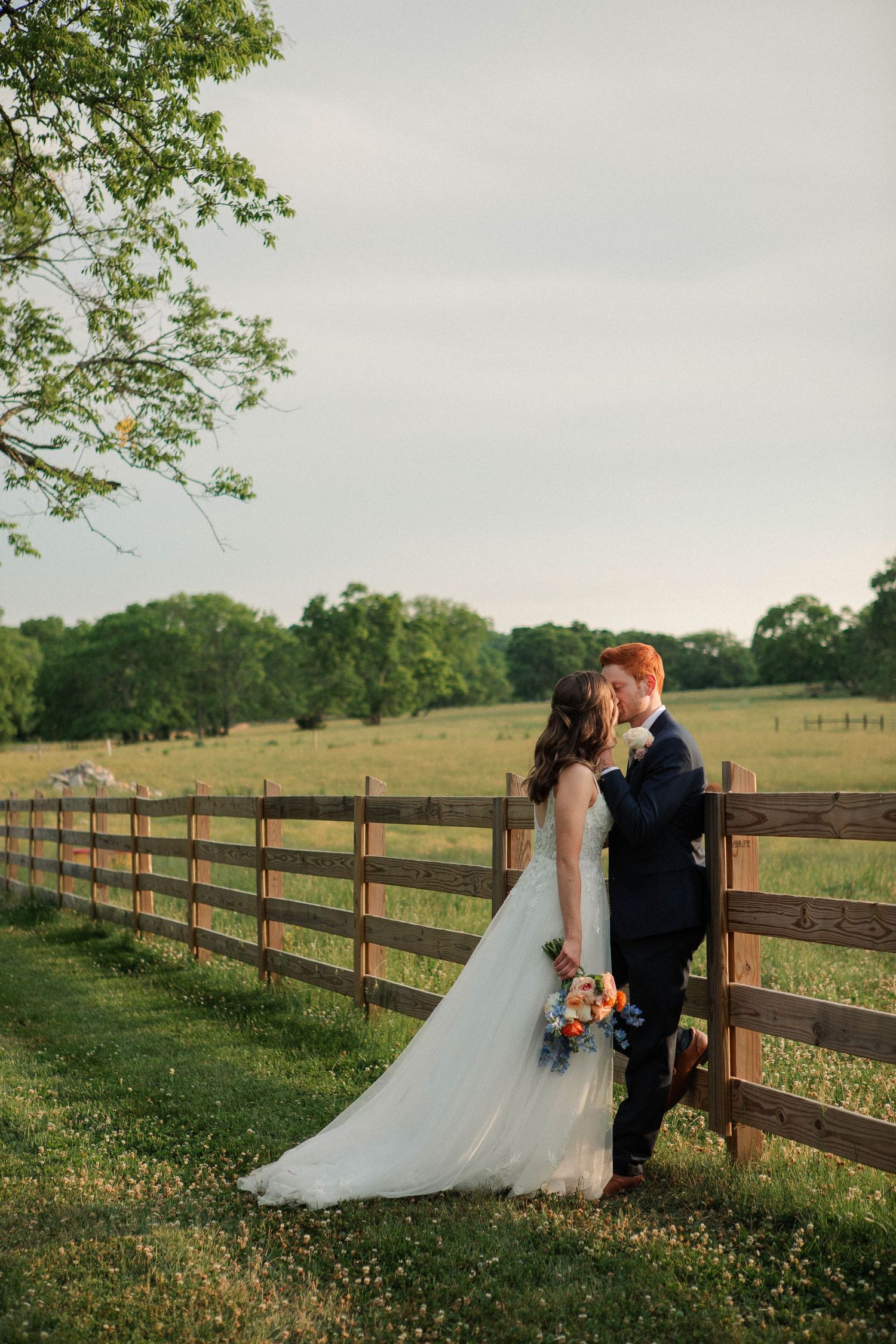 Bride and groom share a kiss beside a rustic wooden fence at golden hour on a lush green farm.