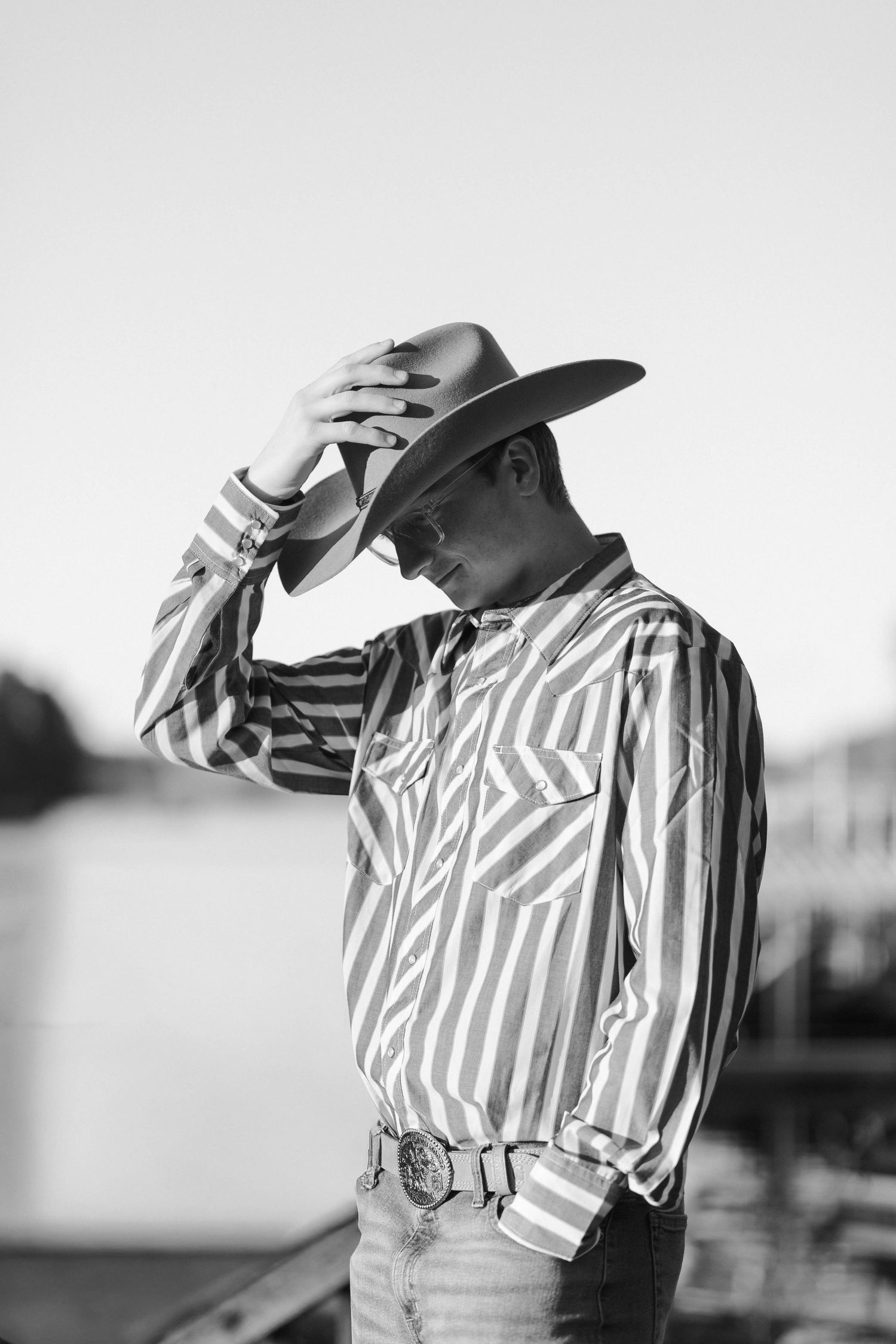 Black and white photo of a young man in striped western shirt and cowboy hat posing outdoors near water.
