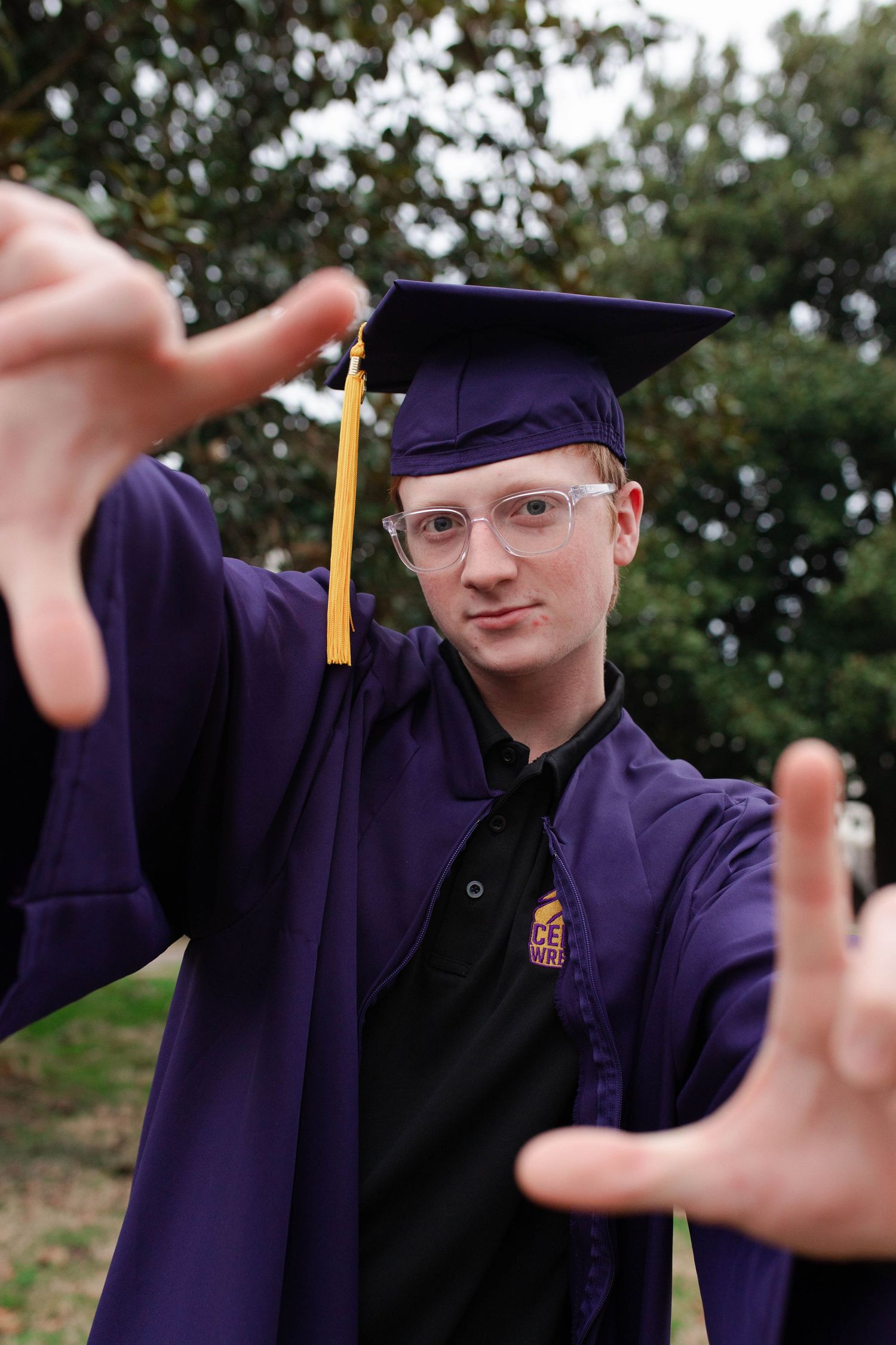 A graduate in purple cap and gown reaches toward camera with both hands, smiling confidently outdoors.