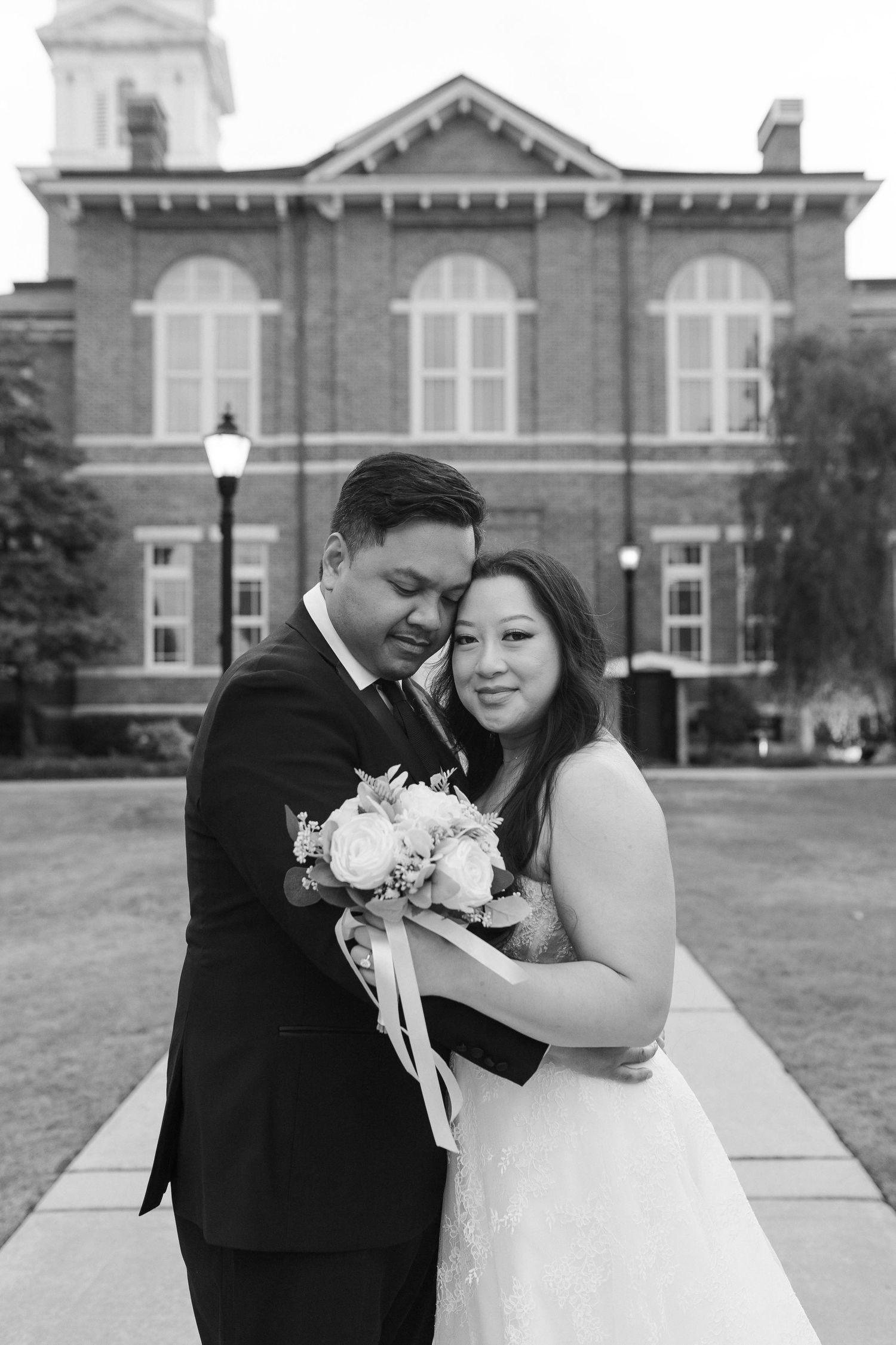 Black and white wedding portrait of bride and groom embracing before historic brick building.
