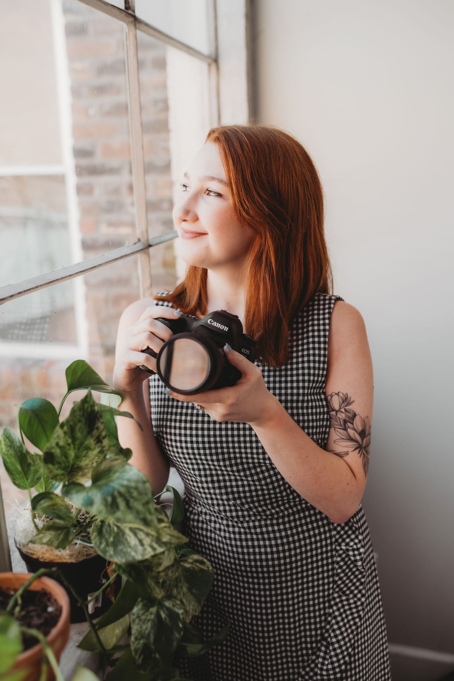 Red-haired woman in gingham dress holding Canon DSLR camera near window with indoor plants and brick wall.