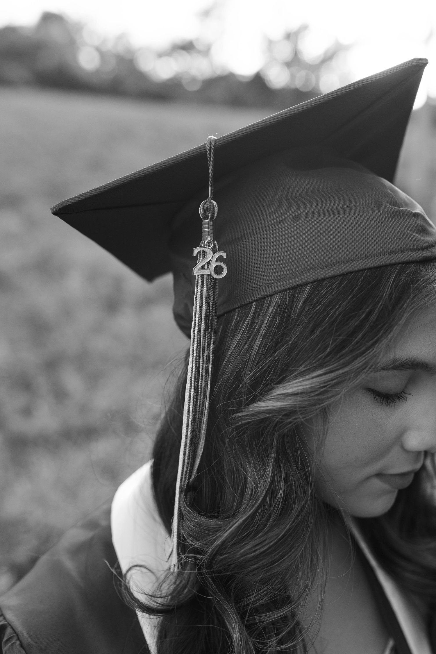 Black and white photo of a graduate wearing a mortarboard cap with a 26 tassel charm, looking down outdoors.