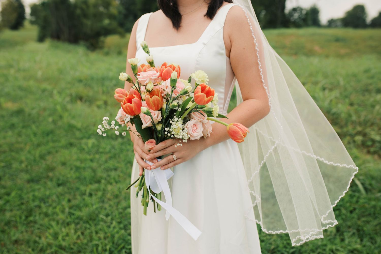 Bride in white dress holding colorful bouquet of coral tulips and pink roses with veil in green field.