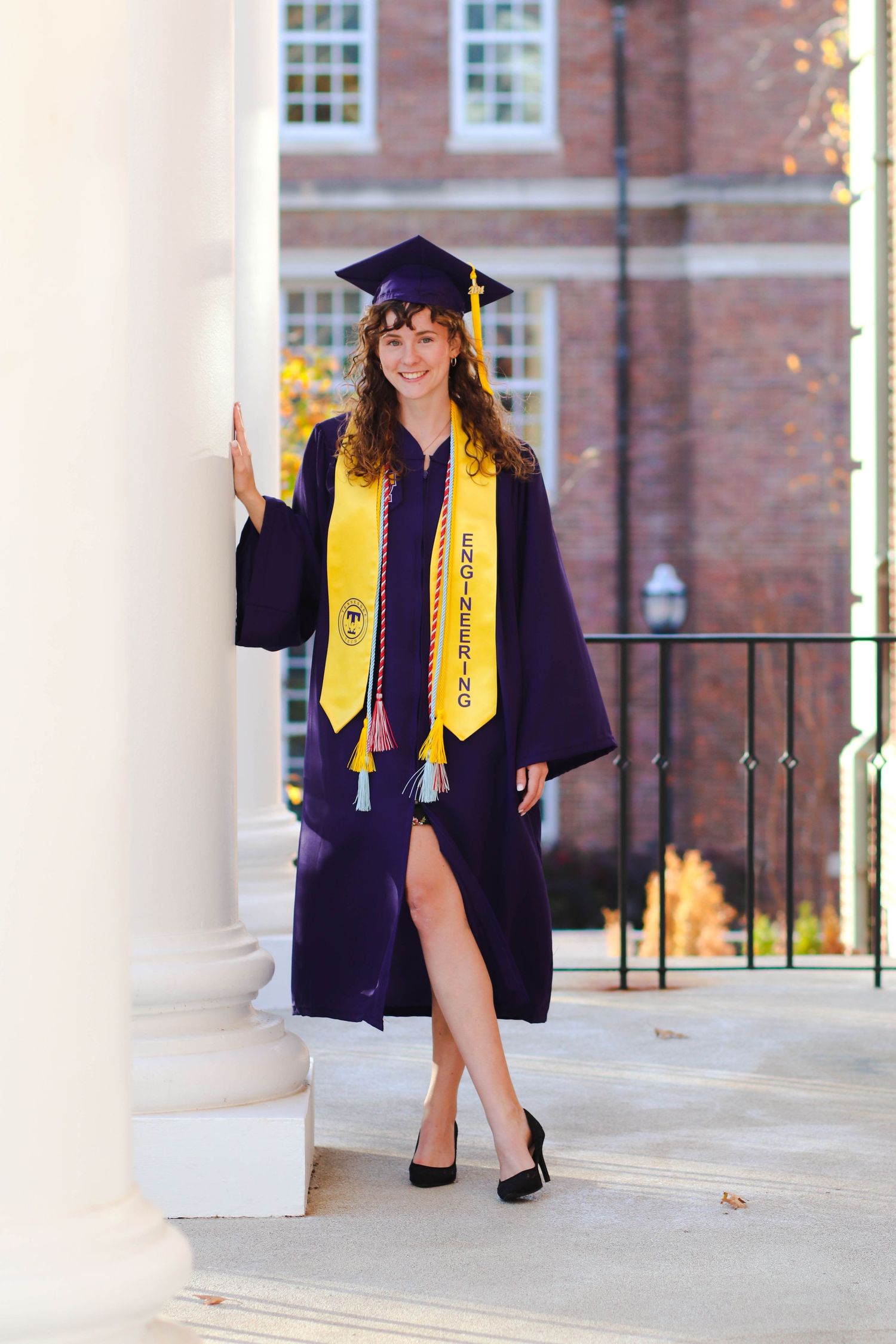 A smiling woman in purple graduation cap and gown with yellow engineering stole poses between white columns at Tennessee Tech.