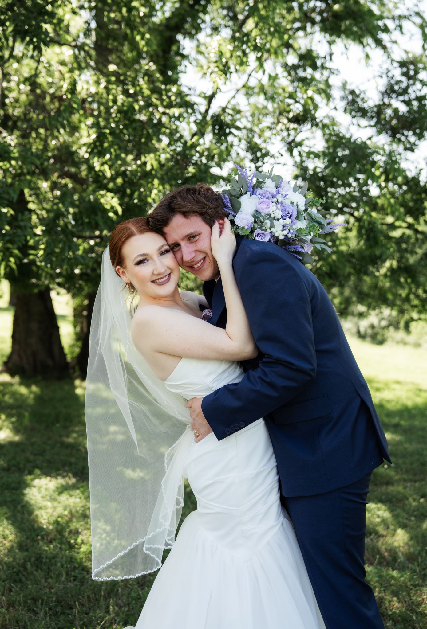 Smiling bride in white gown hugs groom in navy suit while holding purple bouquet under green trees.