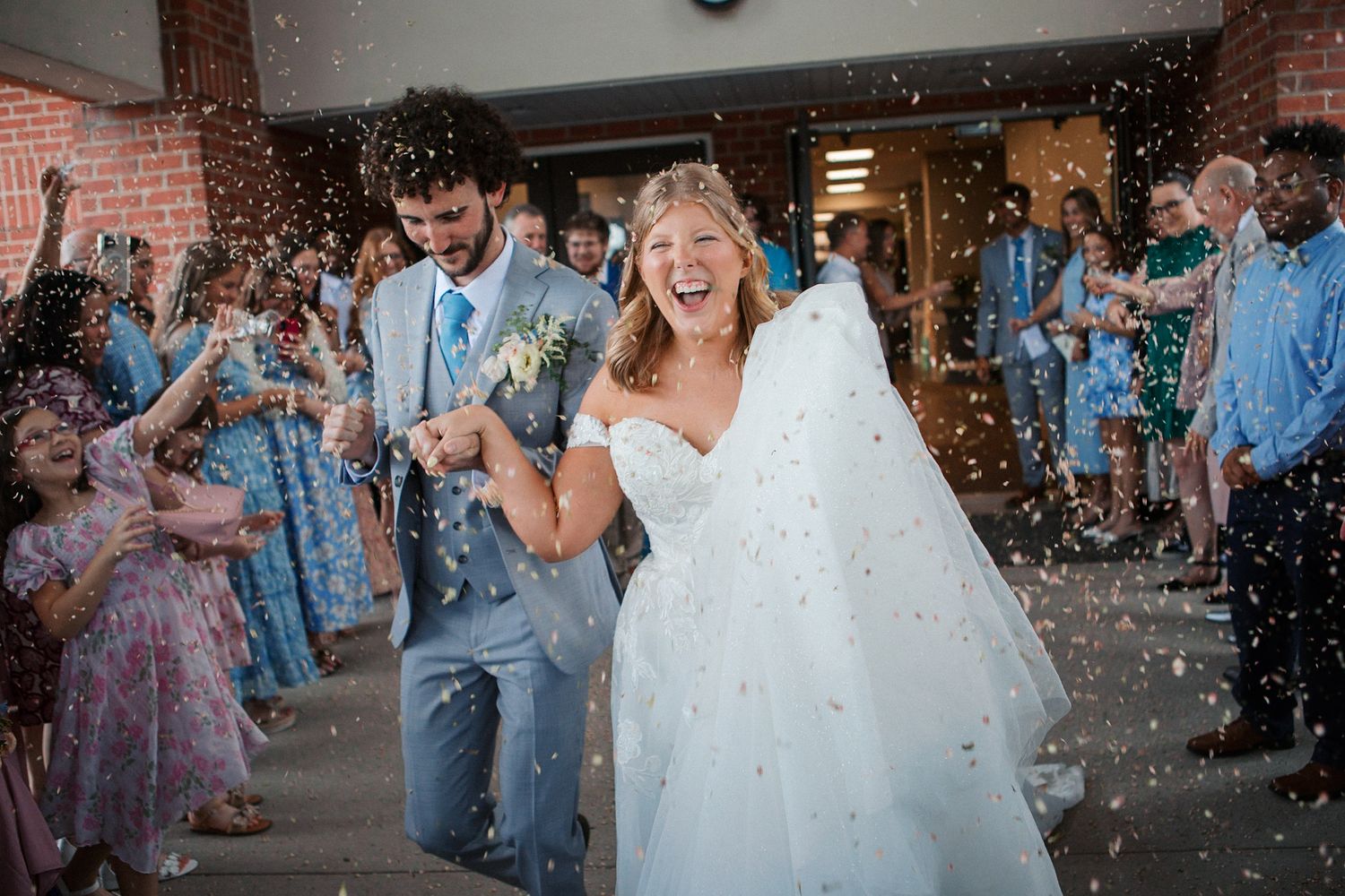 Joyful bride and groom exit church amid confetti shower, guests lining path in blue attire during outdoor wedding celebration.