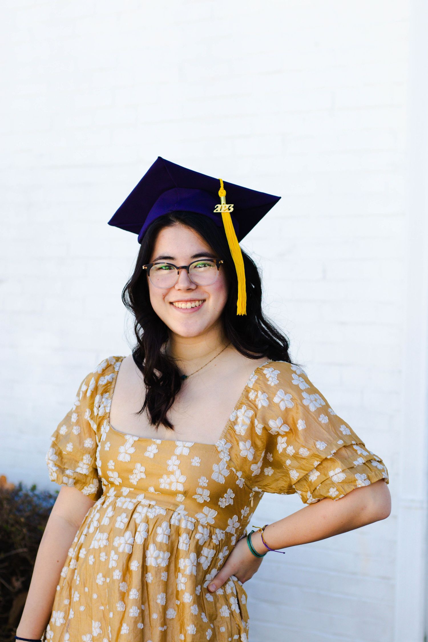 Smiling young woman in yellow floral dress and navy graduation cap with gold tassel posing outdoors.