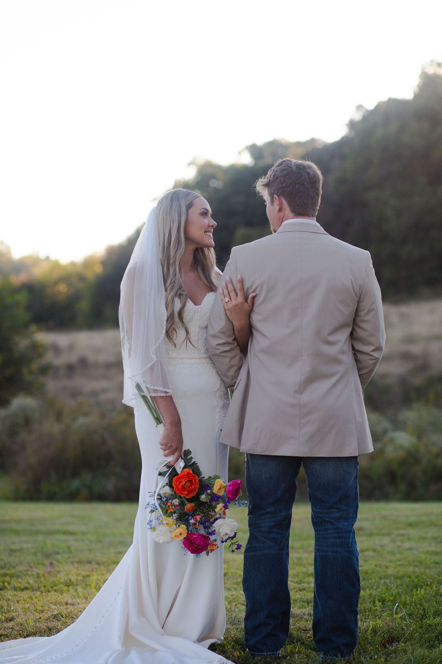 Bride in white gown holding colorful bouquet walks with groom in tan blazer across green field at sunset after eloping in Nashville.