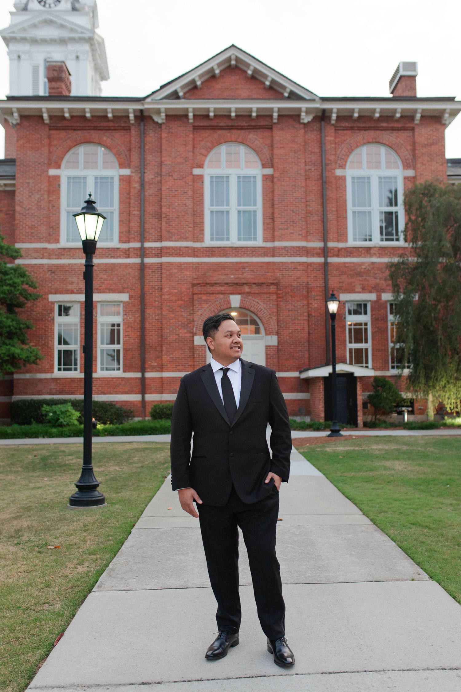 Man in black suit standing on walkway before a historic red brick building with lamp posts.