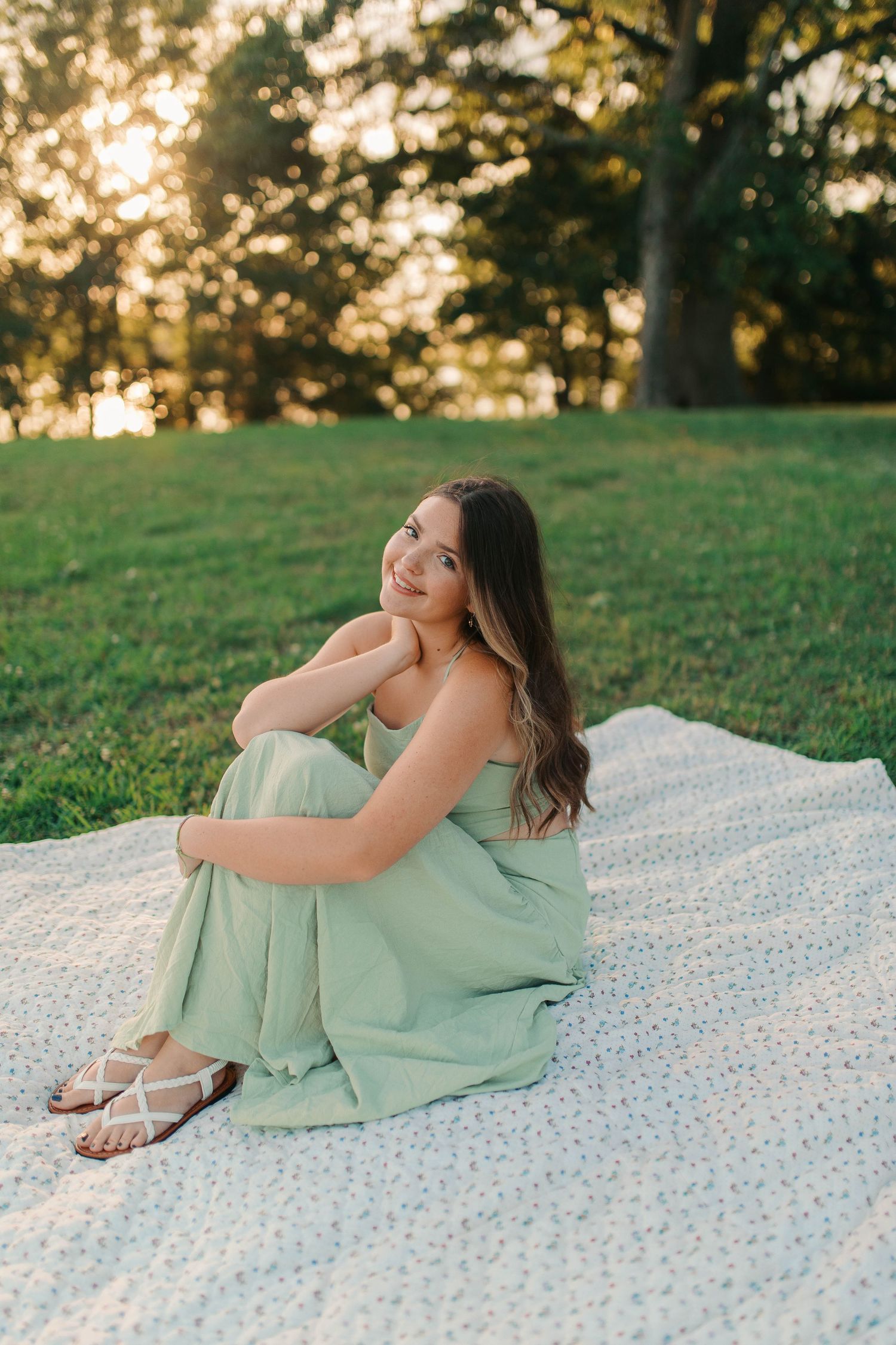 Young woman in mint green dress sitting on blanket outdoors at golden hour sunset in a park.