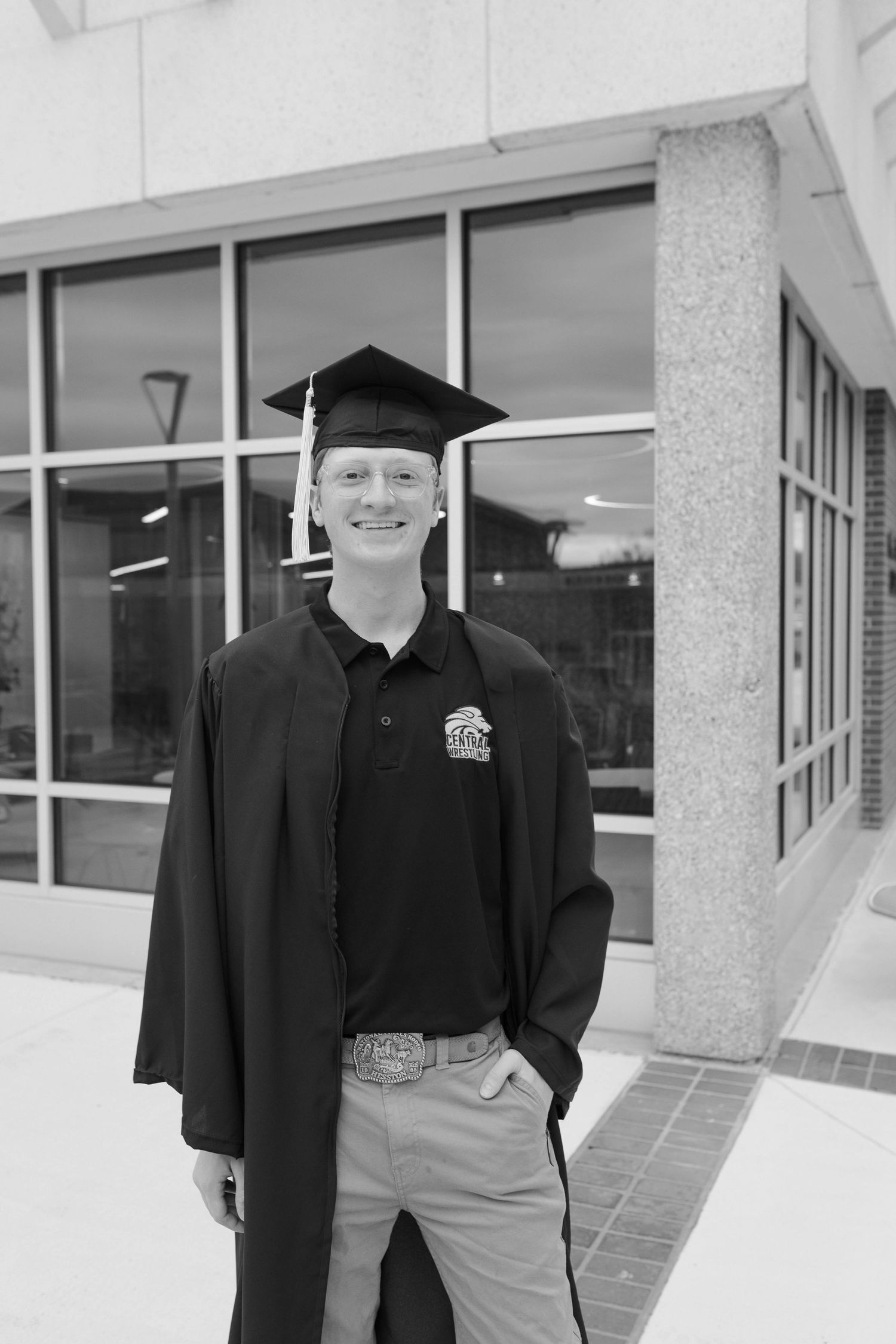 Smiling graduate in cap and gown poses outdoors in black and white photo near a modern building.