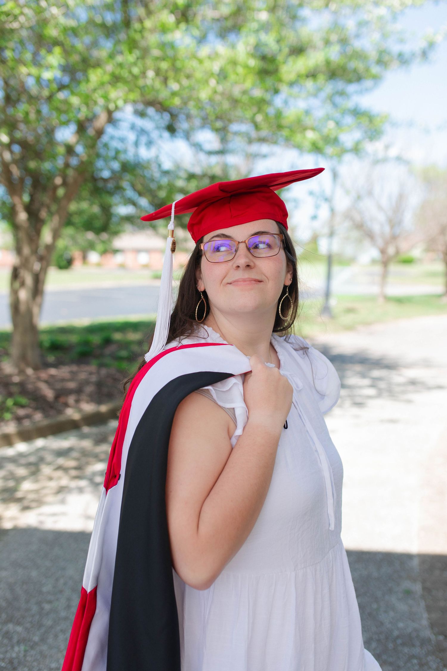 Female graduate in red cap and black gown posing outdoors on sunny graduation day.