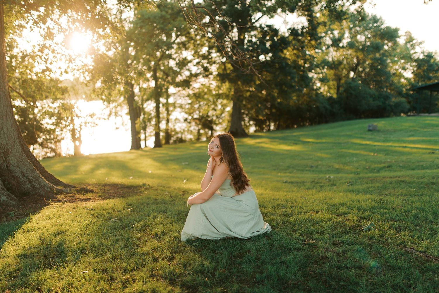Young woman in white dress sitting on lush green grass at golden hour with sunlight filtering through trees.