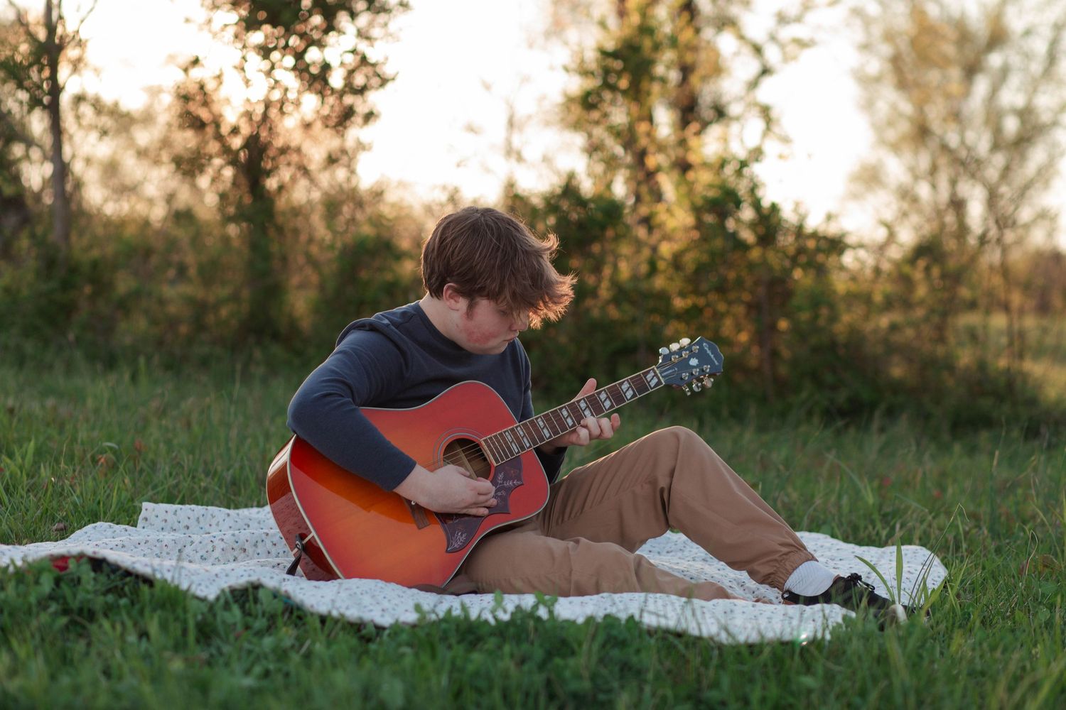 Young musician playing acoustic guitar outdoors on blanket in green field at golden sunset hour.