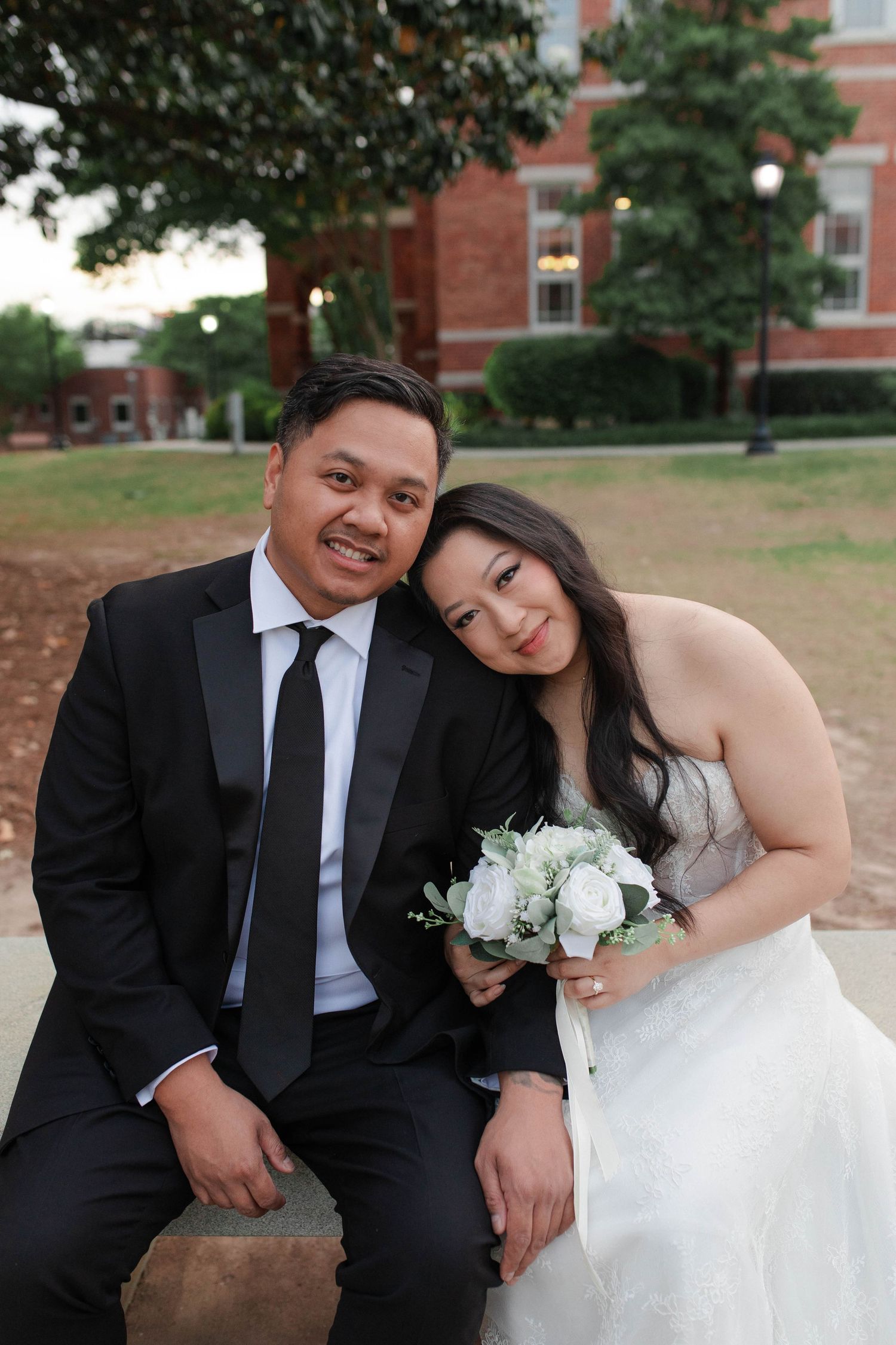 Bride and groom sitting together outdoors, bride holding white bouquet, groom in black tuxedo, brick building background.