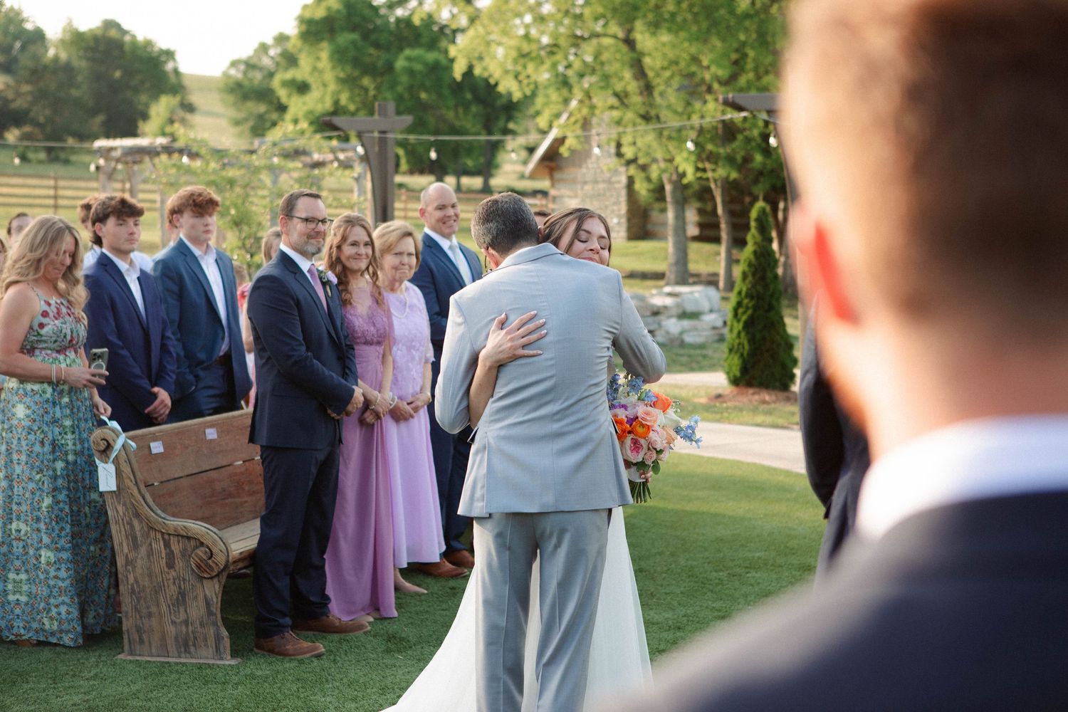 Wedding ceremony outdoors with guests watching as bride and groom embrace on a green lawn with string lights.