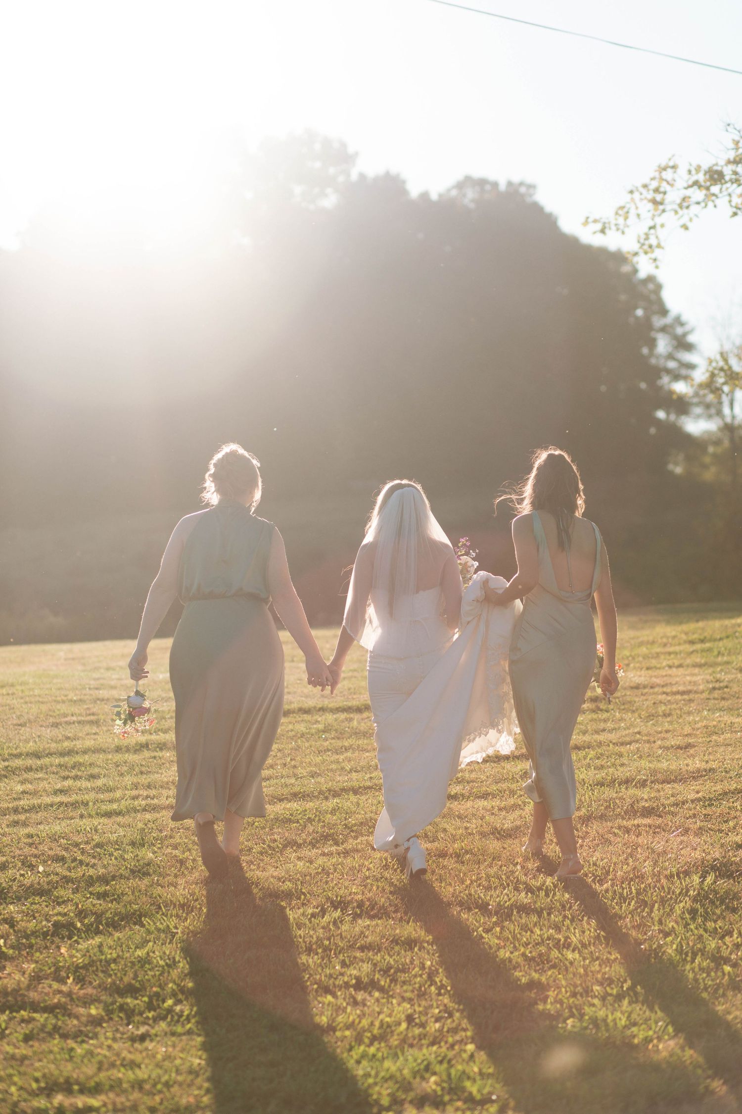 Bride with veil and two bridesmaids walking hand in hand across sunlit grassy field at golden hour after her Nashville elopement.