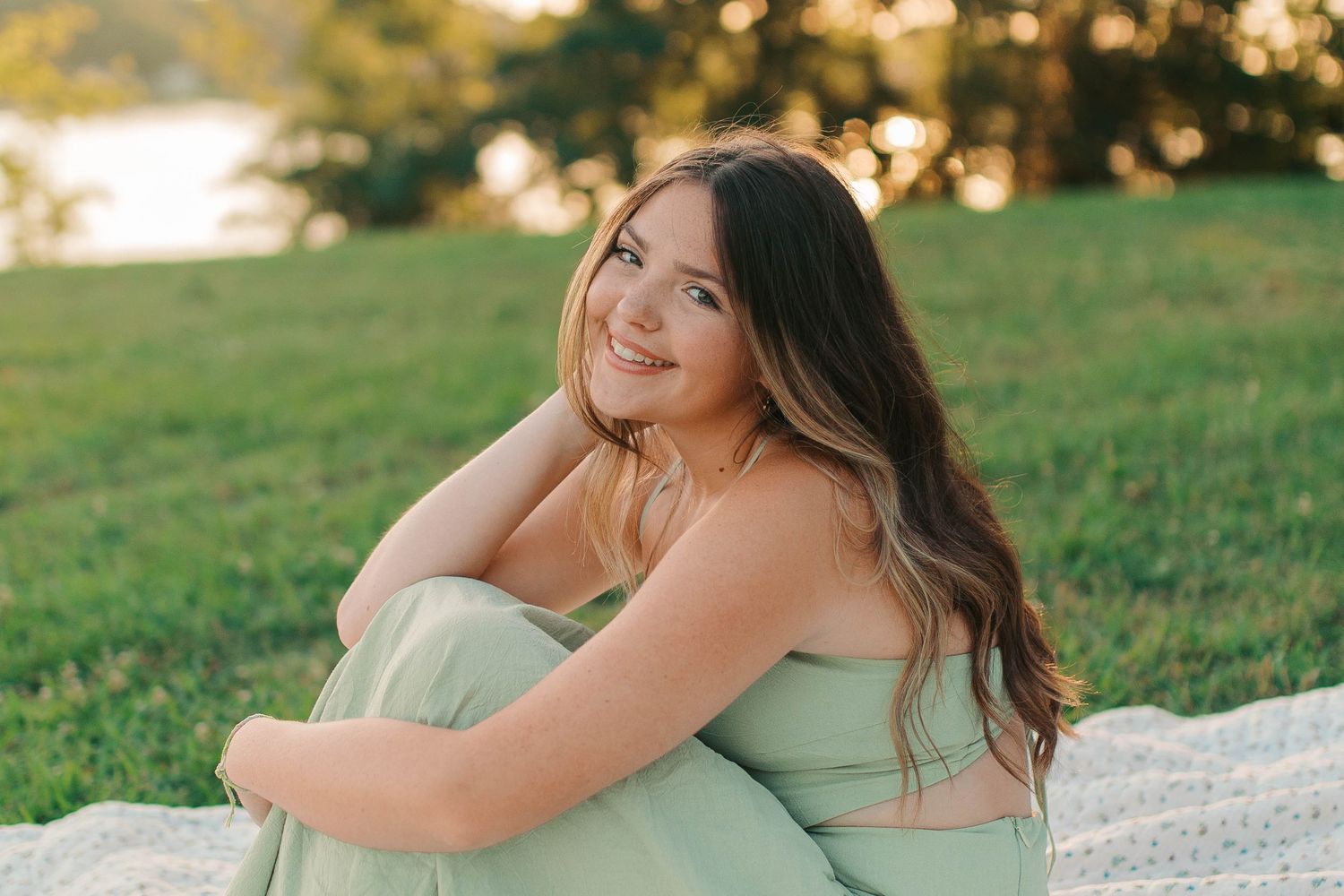Young woman in mint green outfit sitting on grass, smiling over shoulder during golden hour outdoor photo session.