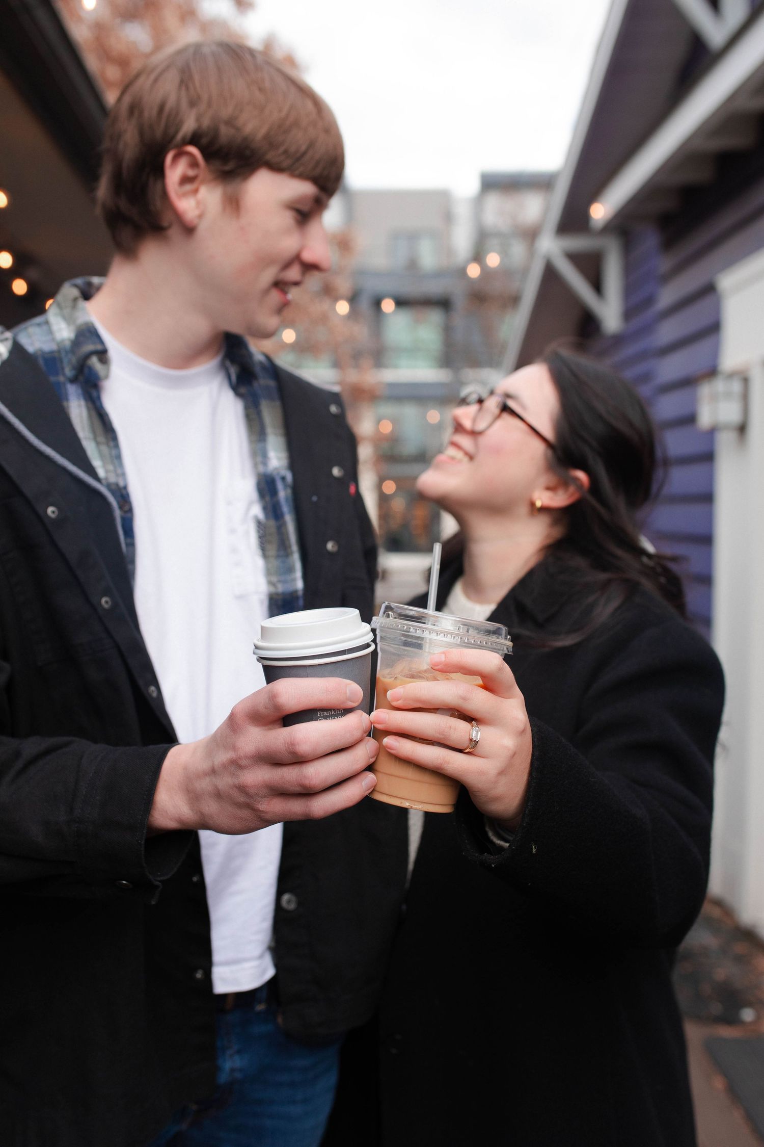 A couple smiling at each other while clinking coffee cups outdoors, with string lights in the background.