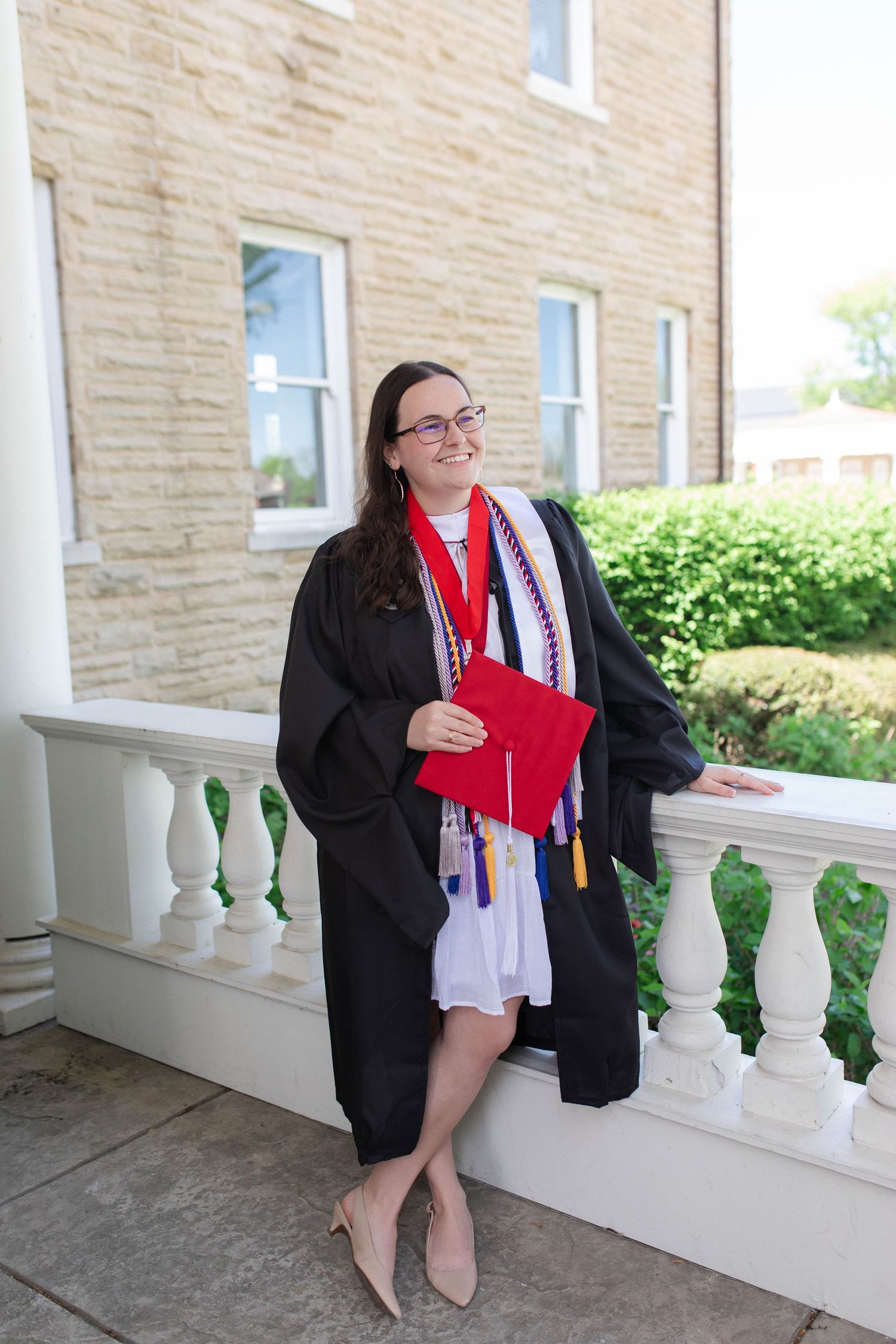Female graduate in black gown holding red diploma cover, leaning on white railing outside brick building.