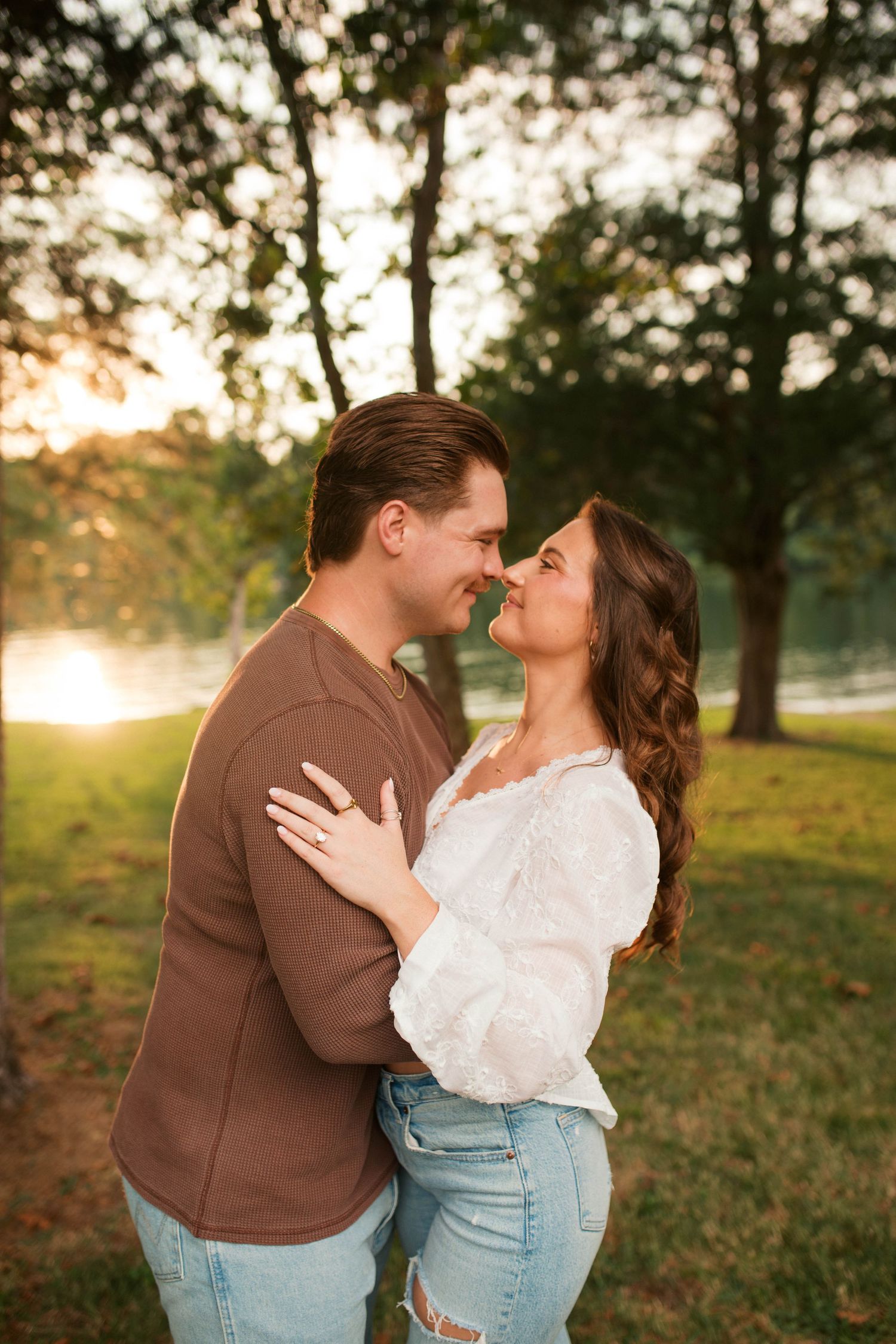 Couple sharing a romantic moment at sunset in a lush green park, woman in white blouse and man in brown sweater.