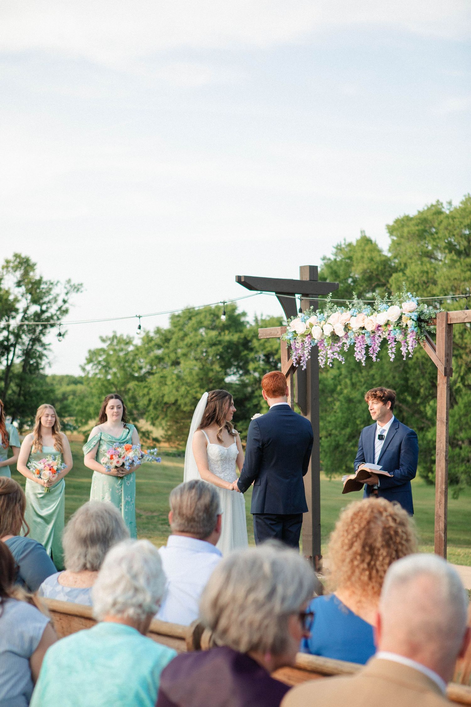 Outdoor wedding ceremony with bride and groom at floral arch, bridesmaids in sage green, and guests seated on a sunny day.